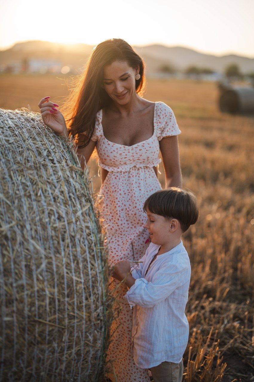 Mama z synkiem na polu przy słomie w promieniach zachodzącego słońca, kobieta w kwiecistej sukience, chłopiec w lnianej koszuli