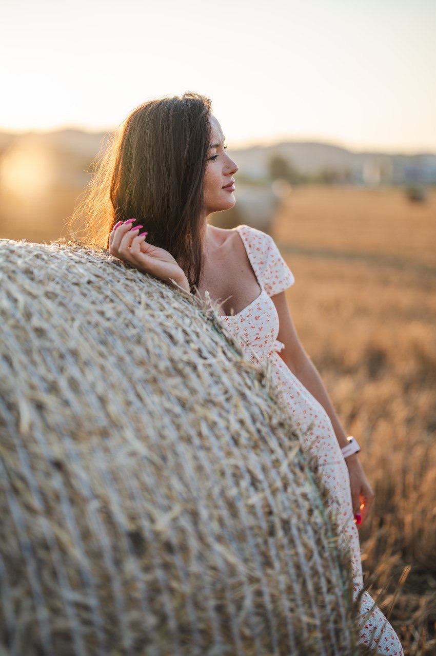 Kobieta w kwiecistej sukience opiera się o belę słomy na polu w promieniach zachodzącego słońca, patrząc w dal.