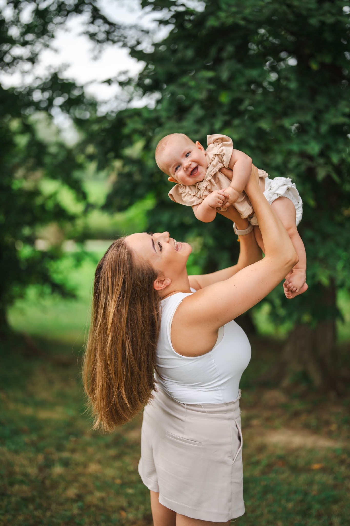 Uśmiechnięta matka w białym topie i beżowych szortach unosi w górę roześmiane niemowlę w beżowej bluzeczce i białych majteczkach na tle zieleni drzew.