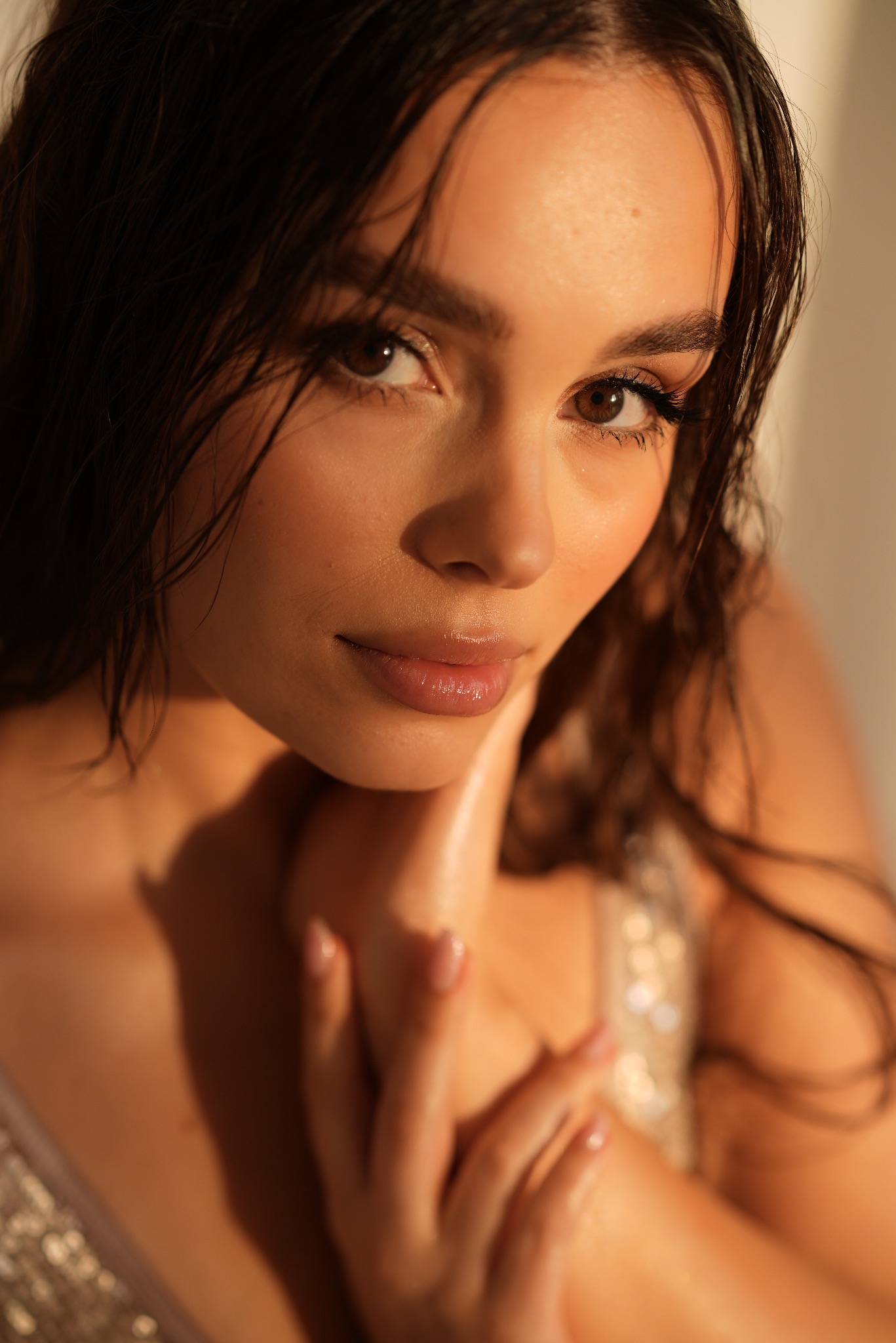 Portret młodej kobiety z mokrymi włosami, delikatnym makijażem i błyszczącą sukienką, oświetlonej ciepłym, naturalnym światłem.