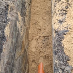 HYDRAULIK Tomasz Krebs - Wykop z piaskiem, fragment pomarańczowej rury kanalizacyjnej i poziomica. Widoczne ślady stóp w piasku. Grunt w wykopie o zróżnicowanej strukturze.