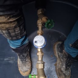 HYDRAULIK Tomasz Krebs - Zamontowany licznik wody z zaworami, widok z góry wewnątrz zbiornika. Obok widoczne buty robocze. Instalacja wodna z mosiężnymi elementami i niebieskimi rurami.