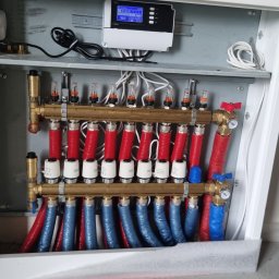 HYDRAULIK Tomasz Krebs - Rozdzielacz ogrzewania podłogowego w szafce, z czerwonymi i niebieskimi rurami, widoczne zawory i elektroniczny sterownik. Instalacja w trakcie montażu, surowa ściana w tle.