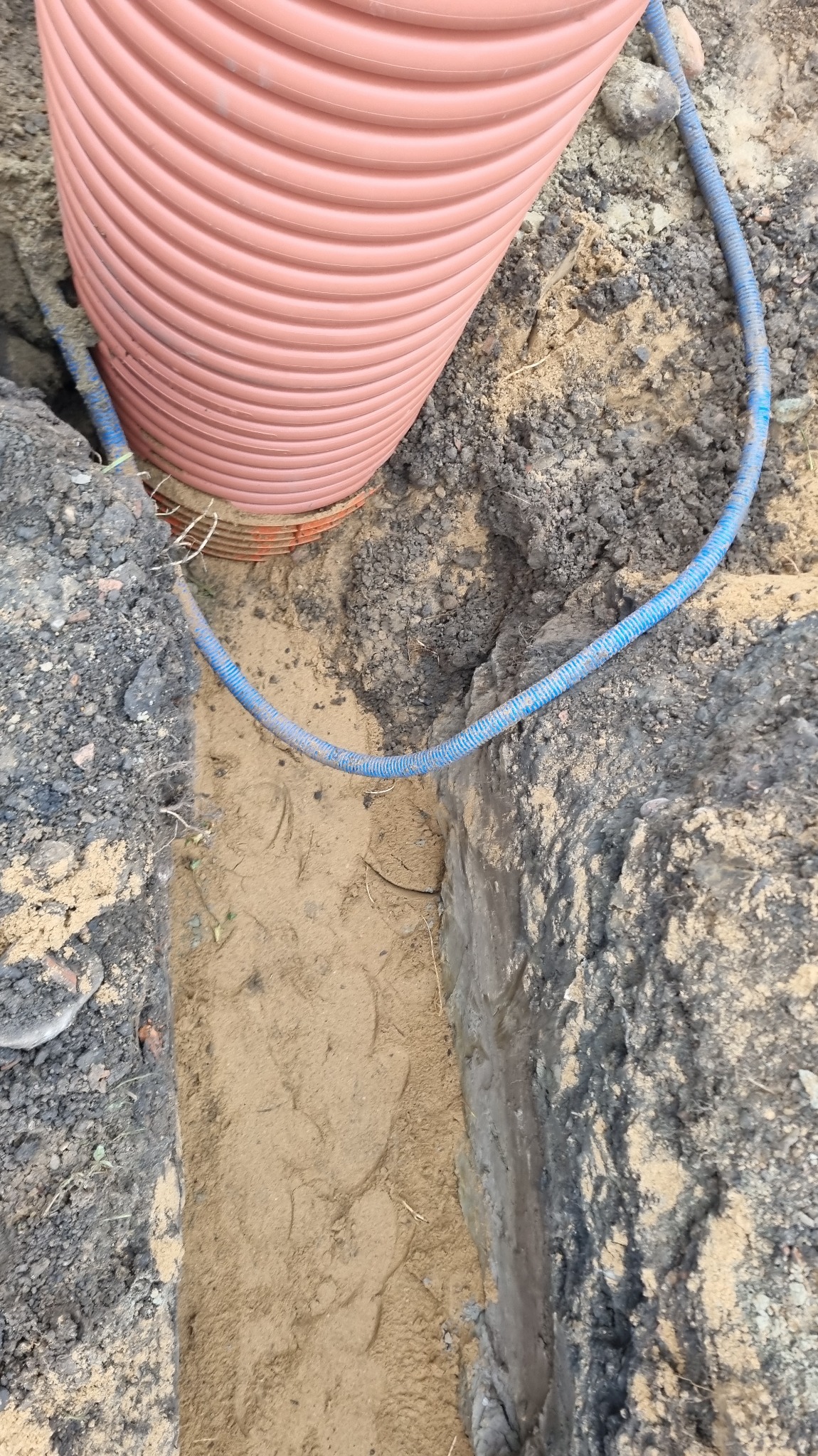 Rura karbowana w wykopie, widoczny fragment niebieskiego przewodu. Prace ziemne w trakcie realizacji, piasek i ziemia.
