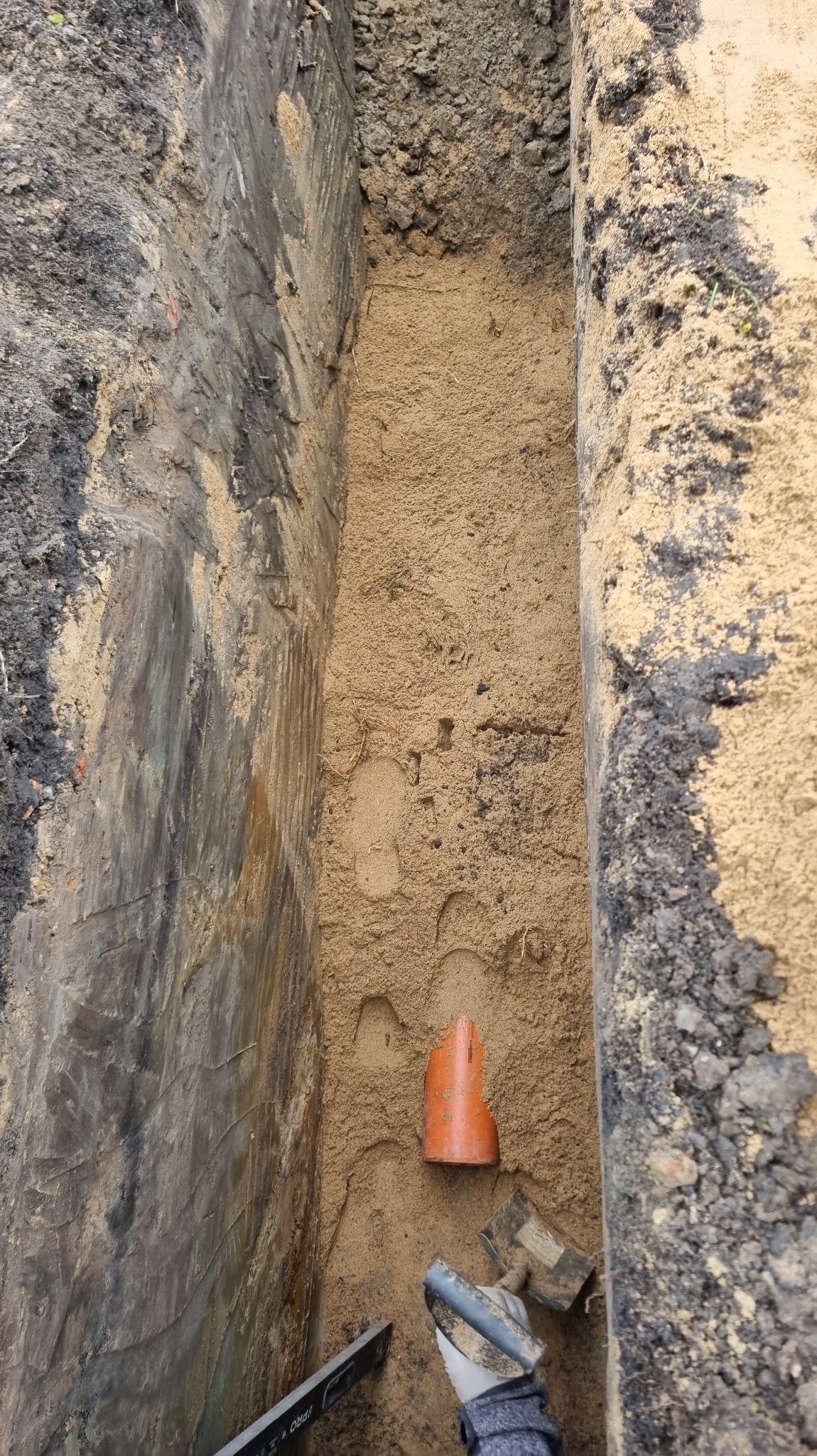 Wykop z piaskiem, fragment pomarańczowej rury kanalizacyjnej i poziomica. Widoczne ślady stóp w piasku. Grunt w wykopie o zróżnicowanej strukturze.