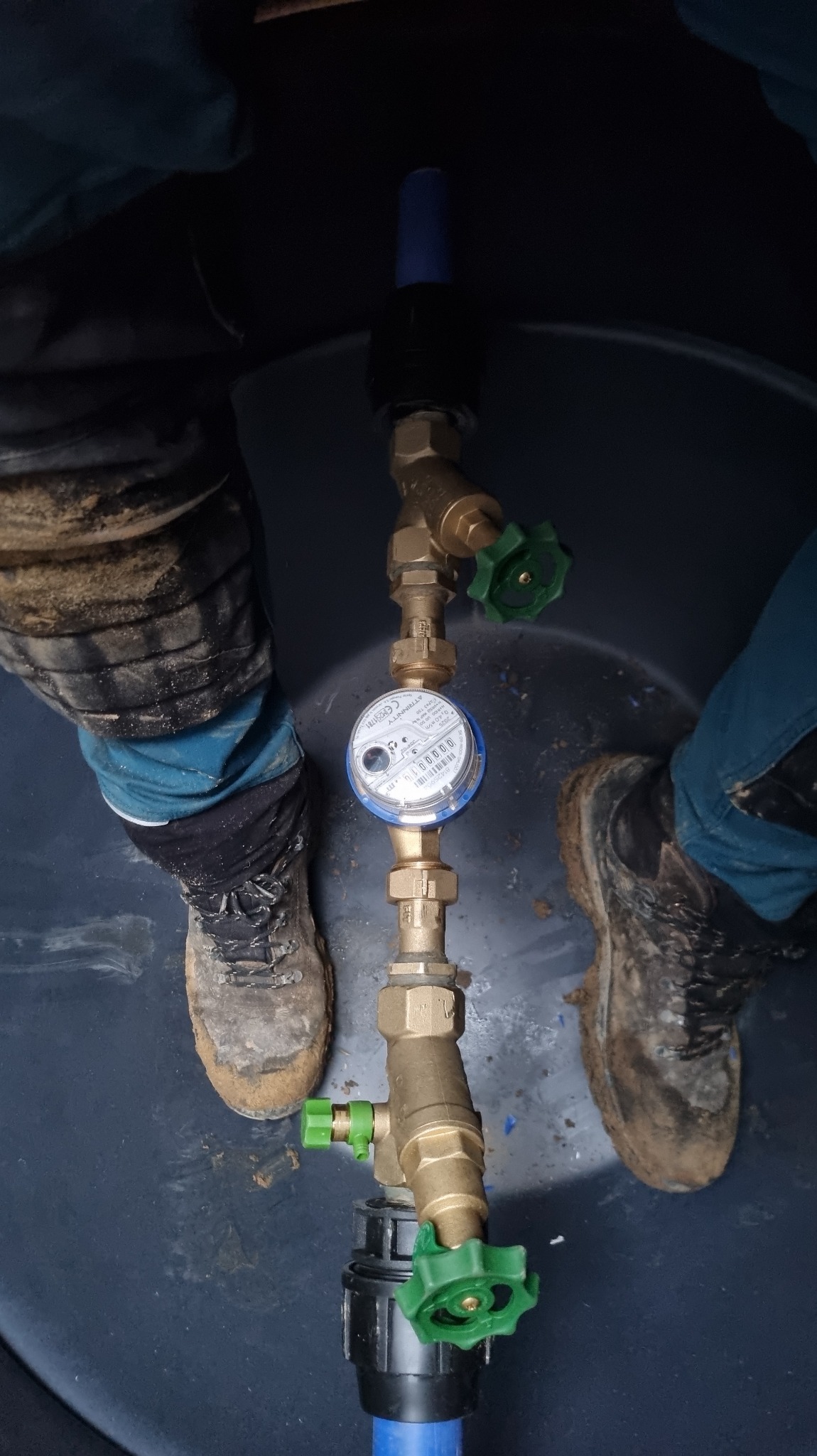 Zamontowany licznik wody z zaworami, widok z góry wewnątrz zbiornika. Obok widoczne buty robocze. Instalacja wodna z mosiężnymi elementami i niebieskimi rurami.