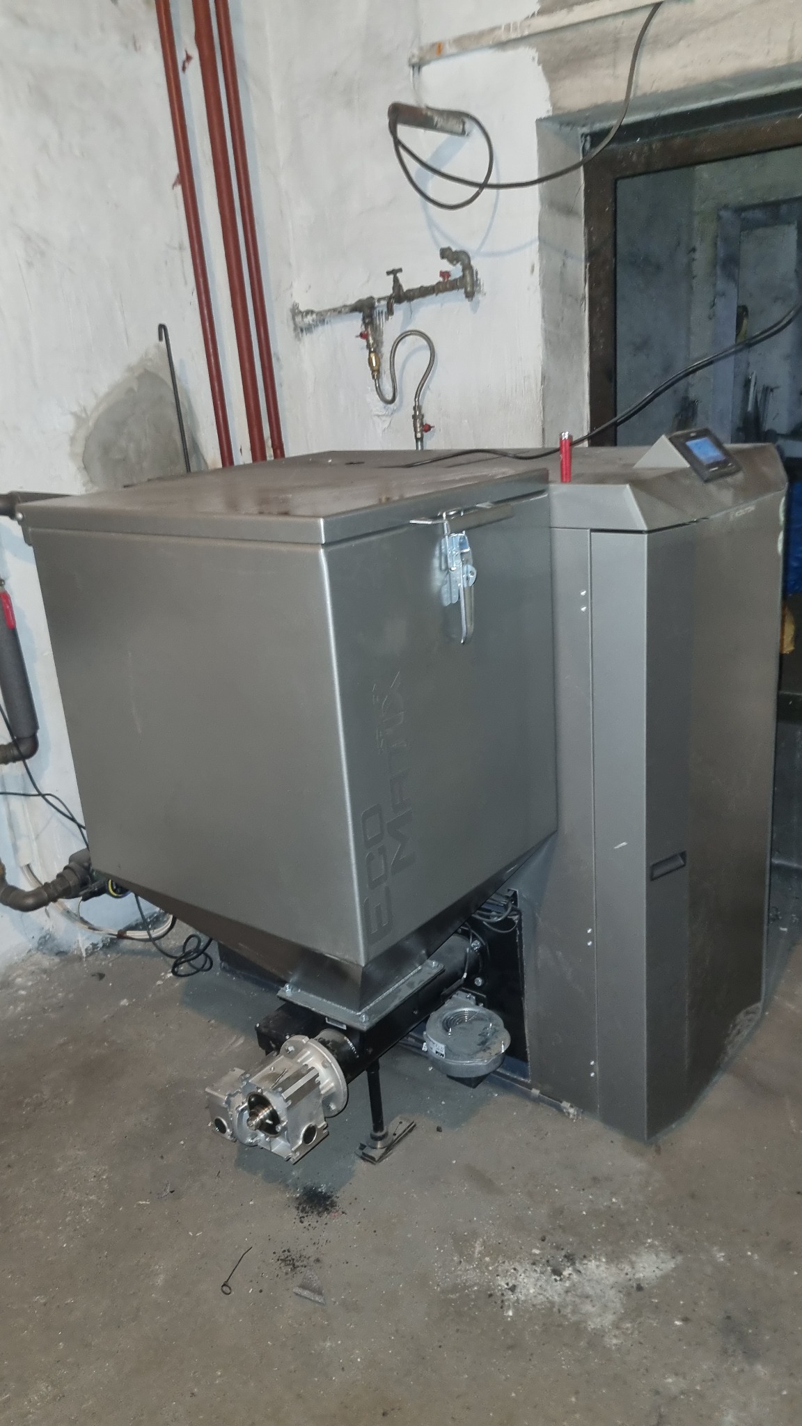 Szary piec na paliwo stałe Eco Matrix w kotłowni, widoczne rury instalacji grzewczej na ścianie. Ujęcie z boku, z bliska, na podłodze widoczne zabrudzenia.