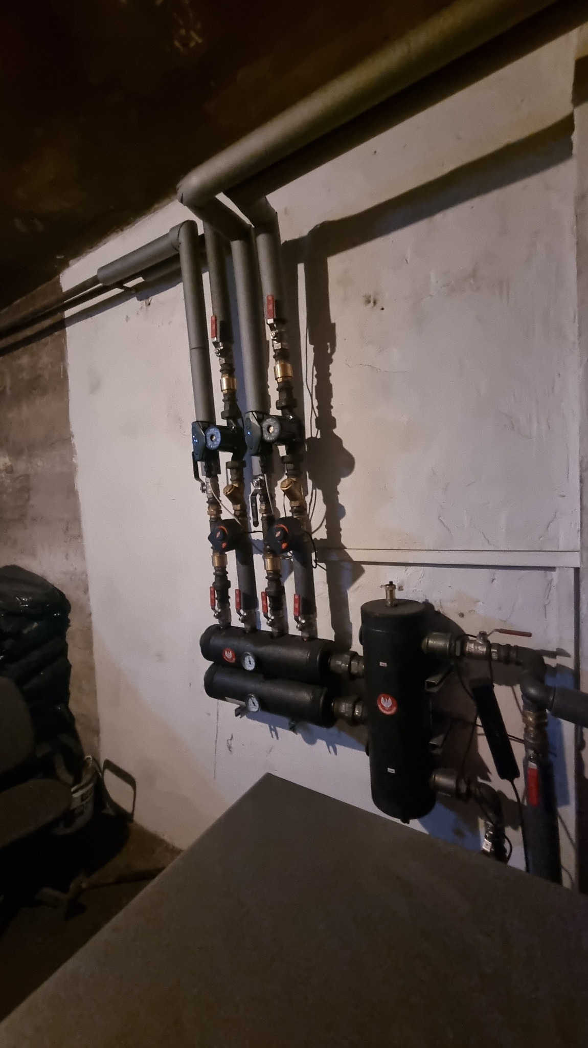 Szara instalacja grzewcza z rurami i zaworami na tle jasnej ściany. Widoczne elementy kotłowni, w tym czarny zbiornik i izolowane rury. Ujęcie z dołu, ciemne oświetlenie.