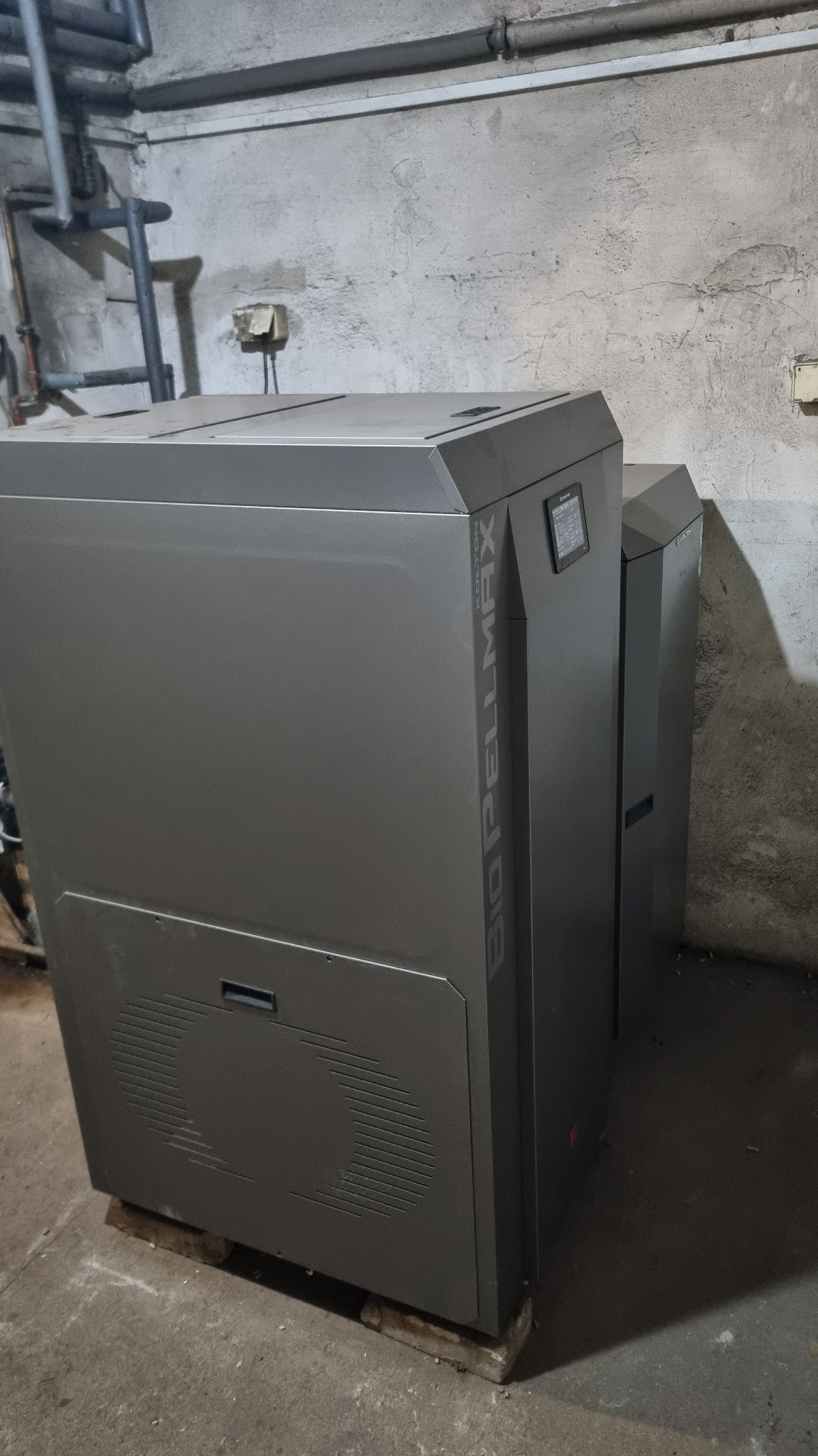 Szary piec na pellet Bio Pellmax w piwnicy, widoczne rury instalacji grzewczej na ścianie. Piec stoi na betonowych bloczkach.