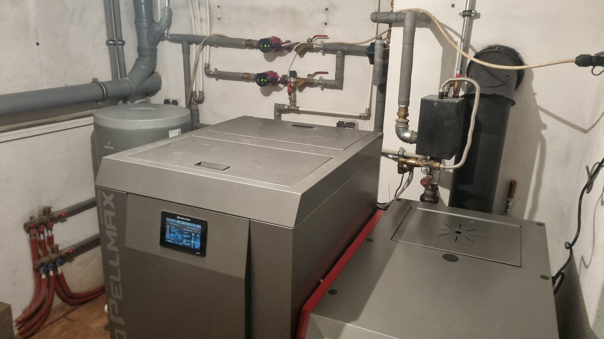 Nowoczesny piec na paliwo stałe Spellmax z ekranem dotykowym, widoczne rury instalacji grzewczej i zbiornik buforowy w kotłowni o białych ścianach.