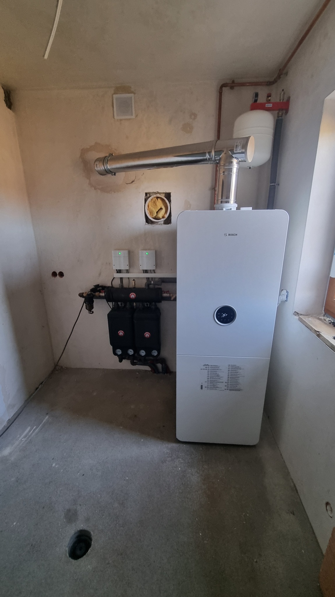 Nowoczesny, biały piec gazowy Bosch z podłączonymi filtrami i rurami w surowym pomieszczeniu. Widoczne elementy instalacji grzewczej i wentylacyjnej.