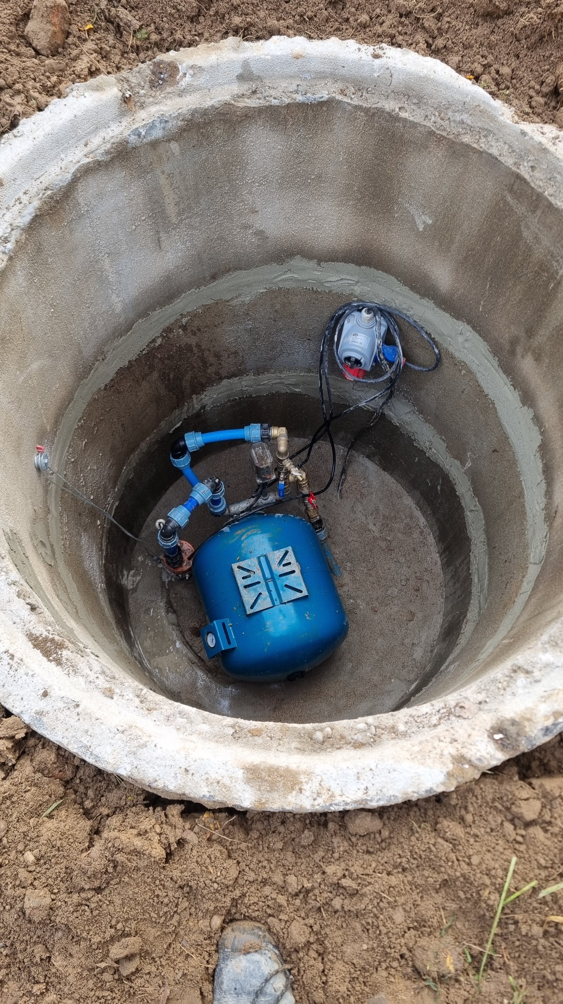 Betonowy krąg studni z niebieskim zbiornikiem hydroforowym i podłączonymi rurami. Widoczny kabel zasilający. Ziemia wokół studni.