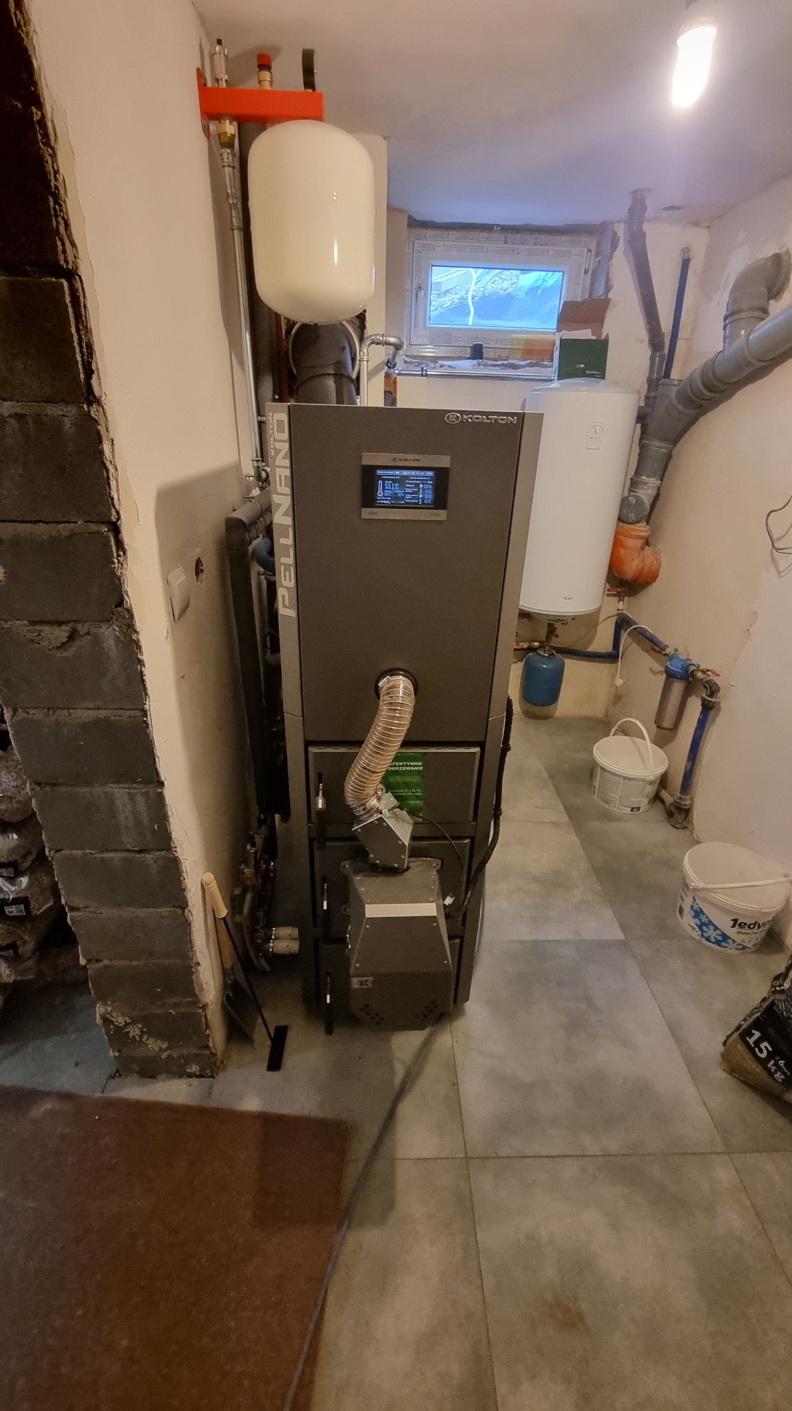 Szary piec na pellet Kolton Pellnano z podajnikiem w kotłowni, widoczne rury, zbiornik wyrównawczy i okno. Ujęcie z dołu, lekko z lewej strony.