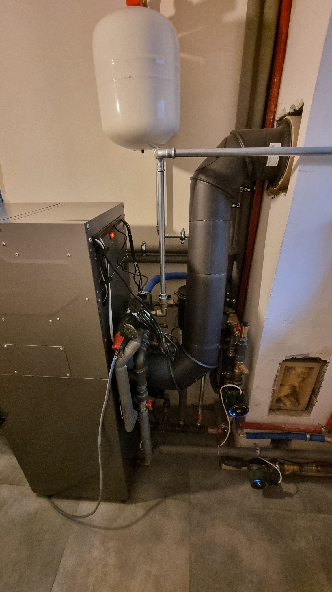 Złożona instalacja grzewcza z piecem na paliwo stałe, zbiornikiem wyrównawczym i rurami odprowadzającymi spaliny. Widoczne liczne zawory i przewody.