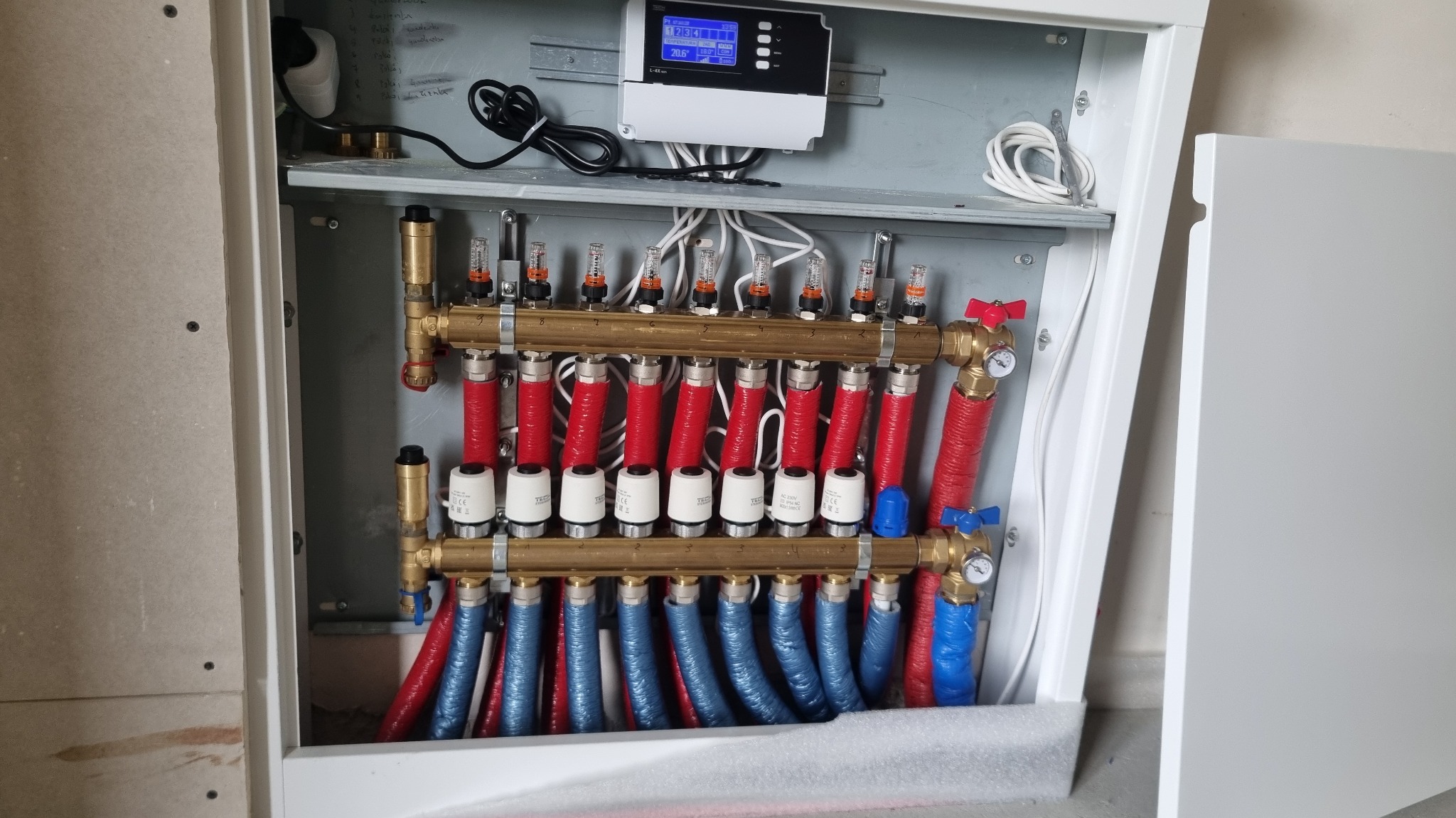 Rozdzielacz ogrzewania podłogowego w szafce, z czerwonymi i niebieskimi rurami, widoczne zawory i elektroniczny sterownik. Instalacja w trakcie montażu, surowa ściana w tle.