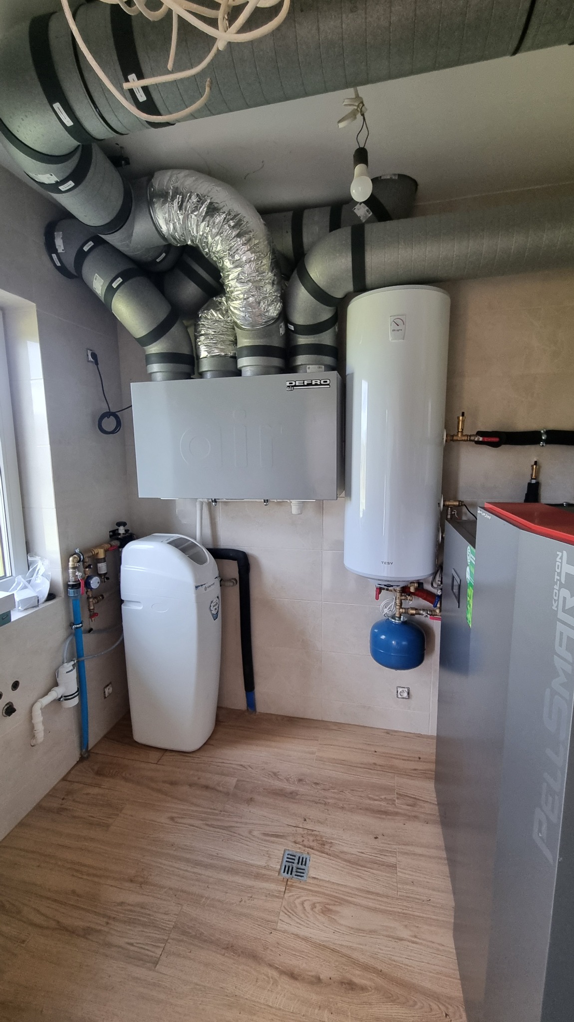 Kotłownia z piecem na pellet, zasobnikiem ciepłej wody i systemem wentylacji. Widoczne rury, filtry i armatura. Podłoga wyłożona panelami imitującymi drewno.