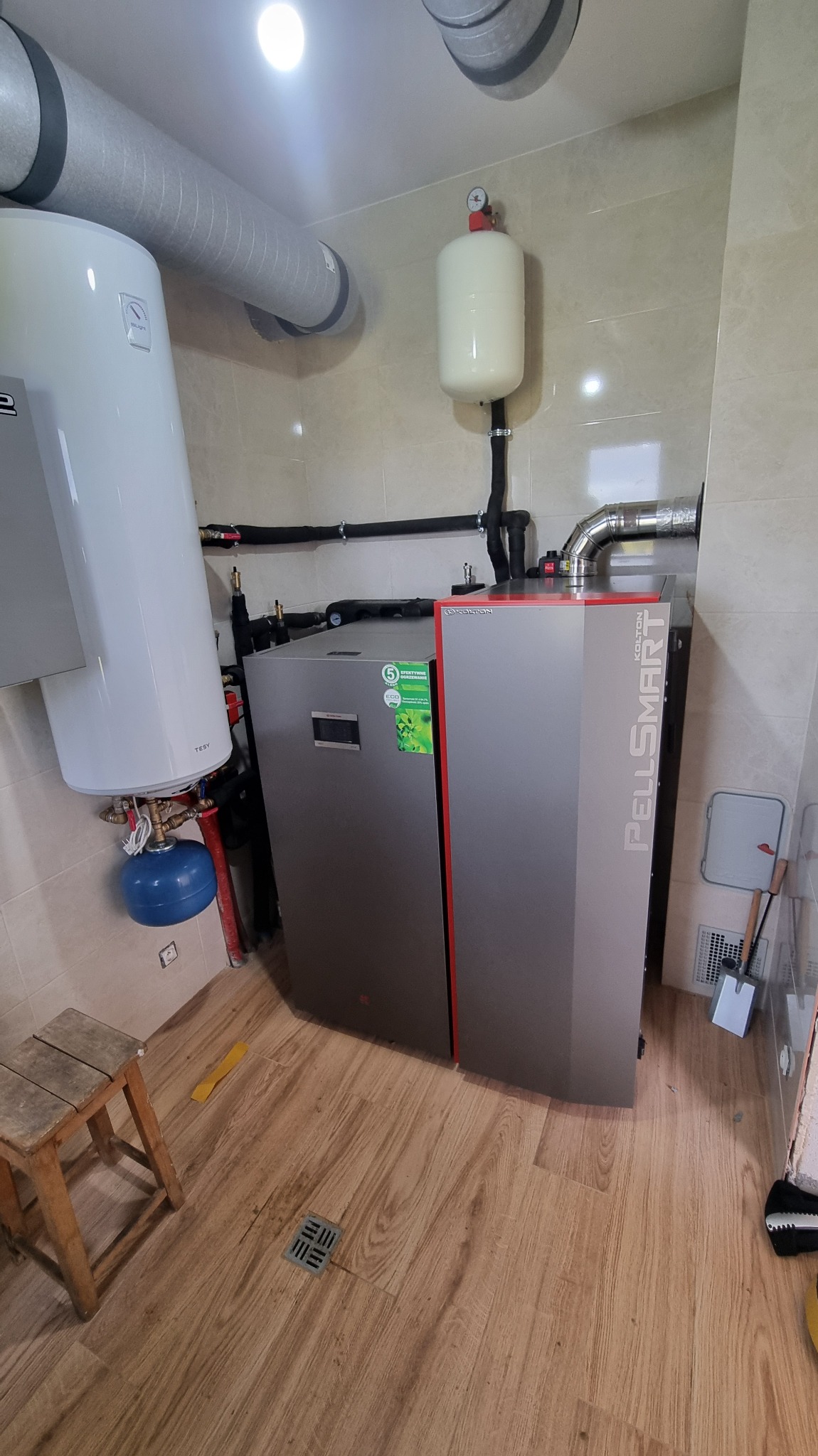 Nowoczesny piec na pellet Kolton PellSmart z osprzętem w kotłowni, widok na instalację, rury i zbiornik wyrównawczy. Drewniana podłoga i białe ściany.