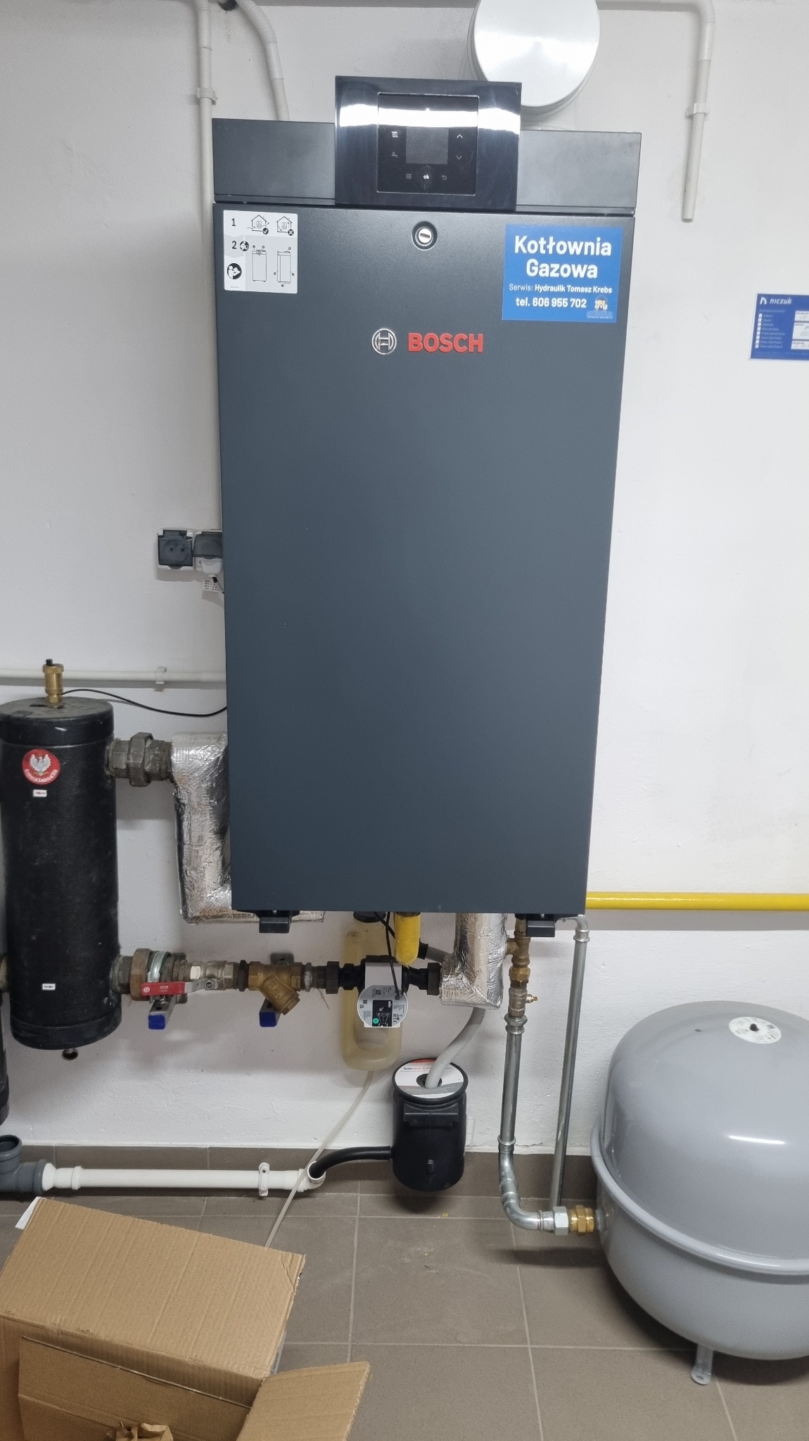 Nowoczesny, szary piec gazowy Bosch z podłączeniami hydraulicznymi i gazowymi w kotłowni. Widoczny zbiornik wyrównawczy i elementy instalacji.