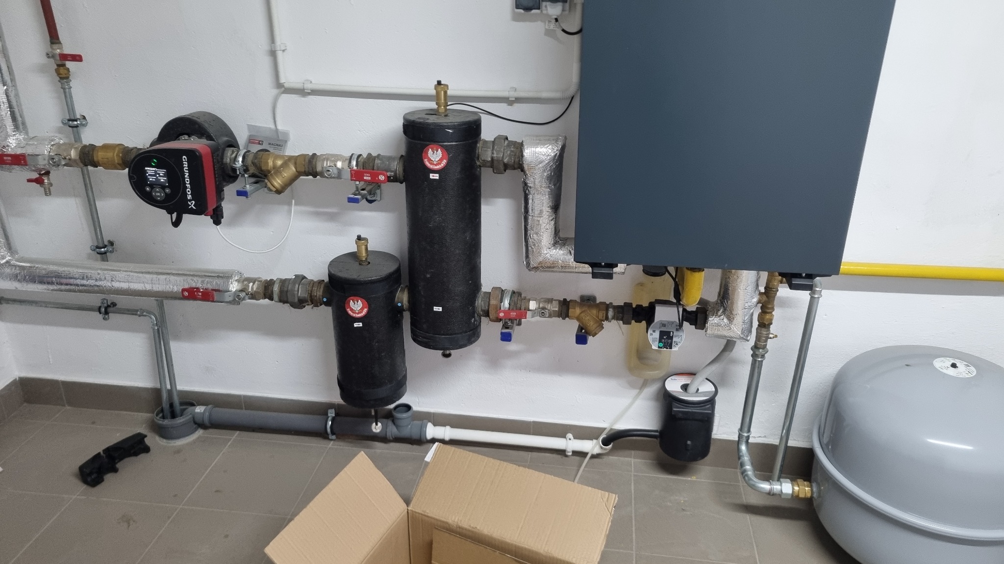 Złożona instalacja grzewcza z kotłem, pompami obiegowymi i zbiornikami wyrównawczymi, rurami z izolacją, zamontowana w kotłowni z szarą podłogą.