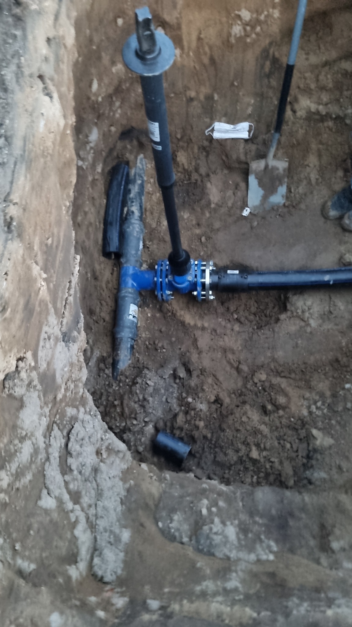 Niebieski trójnik instalacji wodnej w wykopie ziemnym, z widoczną rurą pionową i poziomą. Obok leży maska ochronna i stoi szpadel. Widoczne złącza skręcane.