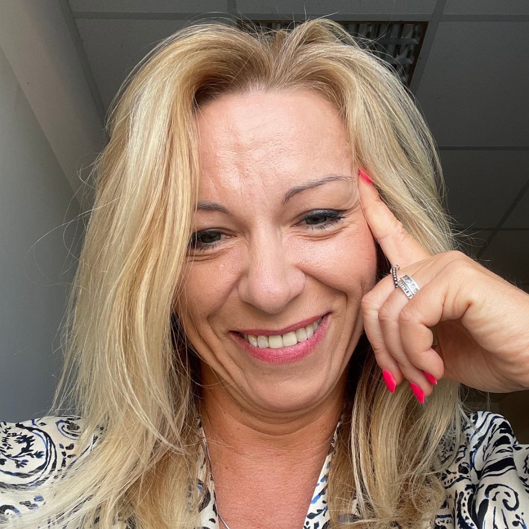 Uśmiechnięta kobieta z blond włosami i pierścionkiem na palcu, dotykająca palcem czoła, w bluzce we wzory, z pomalowanymi na różowo paznokciami.