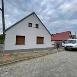 Świeżo pomalowany dom jednorodzinny z brązowymi roletami, widoczny mężczyzna w ubraniach roboczych wychodzący z białego busa na tle brukowanej ulicy.