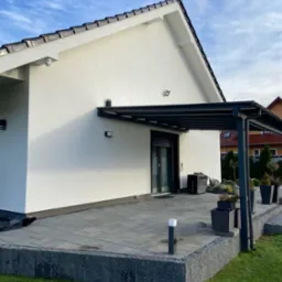 Nowoczesny dom z jasną elewacją, tarasem z szarej kostki brukowej, zadaszeniem i fragmentem trawnika z elementami montażowymi