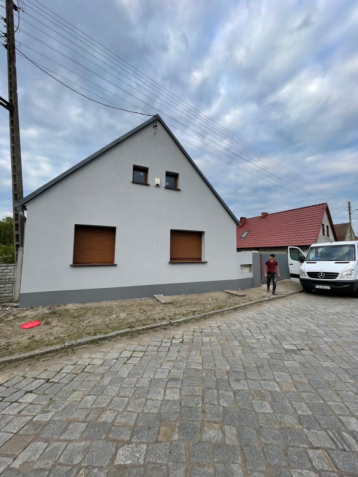 Świeżo pomalowany dom jednorodzinny z brązowymi roletami, widoczny mężczyzna w ubraniach roboczych wychodzący z białego busa na tle brukowanej ulicy.