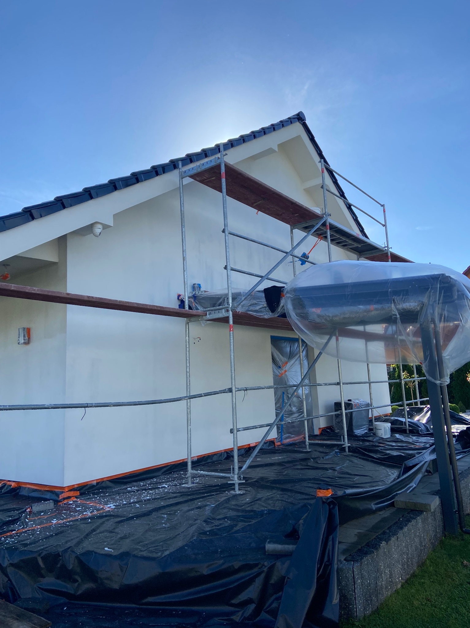 Świeżo pomalowana, biała elewacja domu jednorodzinnego, widoczna ze rusztowania, z zabezpieczonymi oknami i podłożem folią ochronną, w słoneczny dzień.