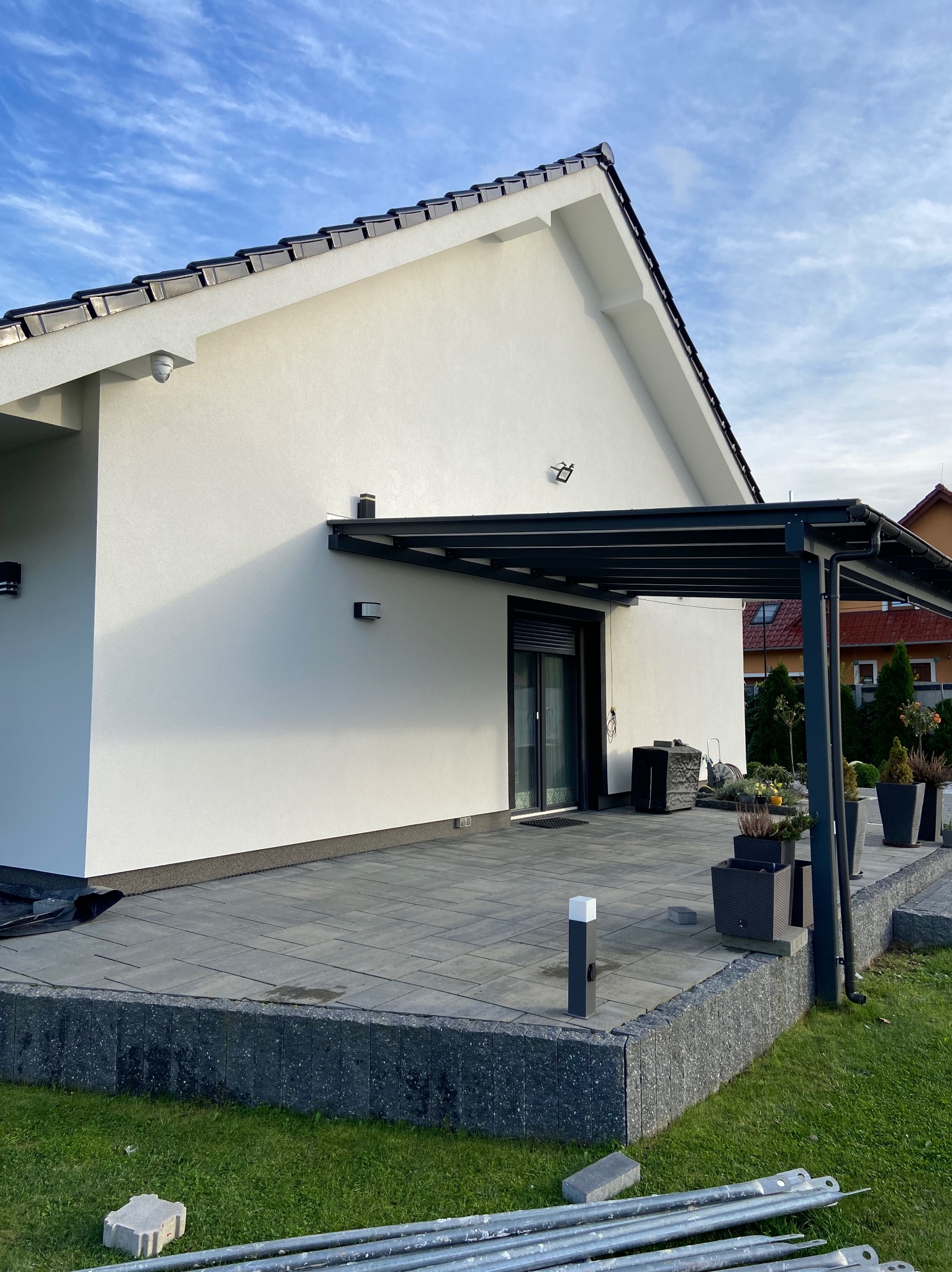 Nowoczesny dom z jasną elewacją, tarasem z szarej kostki brukowej, zadaszeniem i fragmentem trawnika z elementami montażowymi