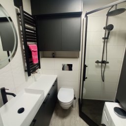 PD BUD - Nowoczesna łazienka z czarnymi akcentami: armatura, grzejnik drabinkowy z różowym ręcznikiem, kabina prysznicowa typu walk-in z deszczownicą i szafki wiszące nad toaletą.