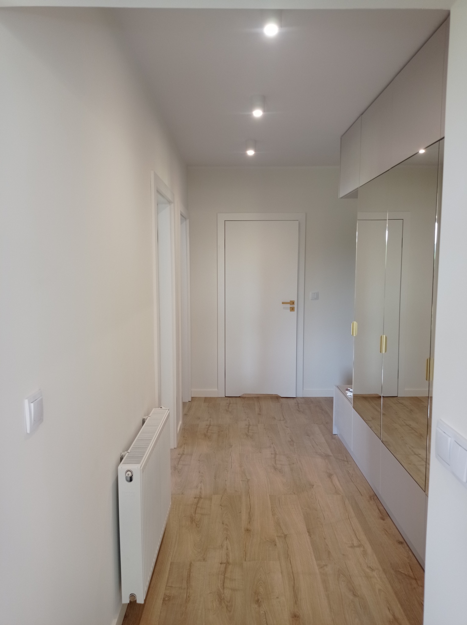 Jasny korytarz po remoncie z panelami podłogowymi, grzejnikiem, białymi drzwiami z złotymi klamkami i szafą z lustrzanymi frontami.