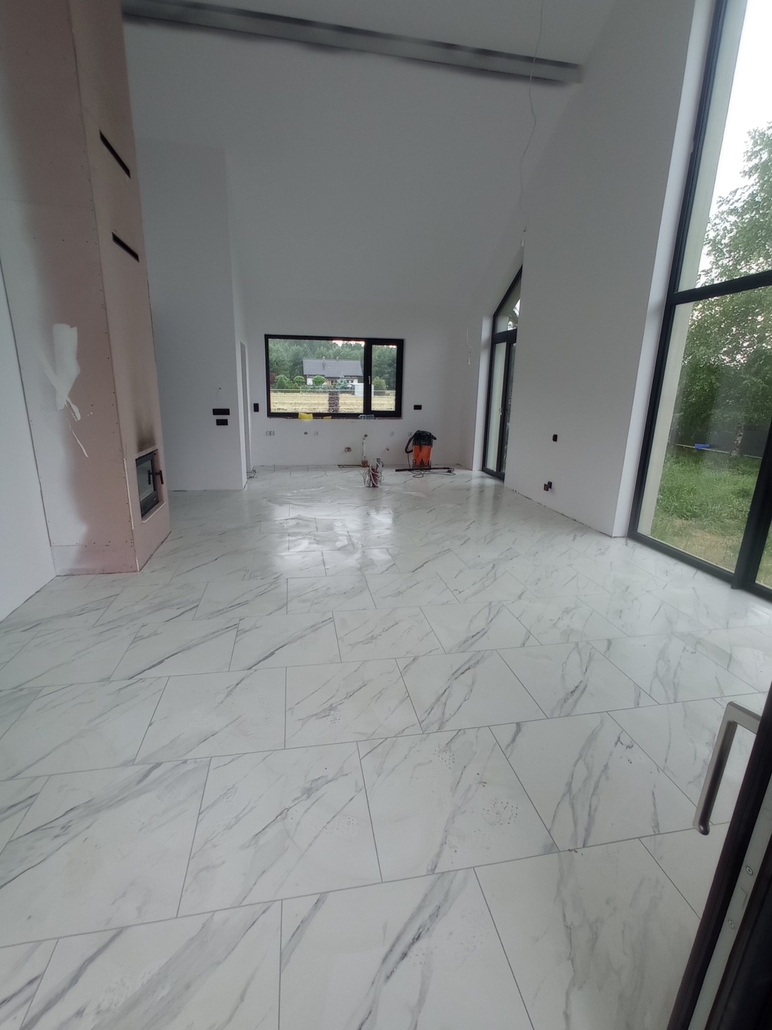 Przestronne wnętrze w trakcie prac wykończeniowych, z ułożoną białą podłogą imitującą marmur, widoczne ślady po szpachlowaniu na obudowie kominka oraz pracownik w pomarańczowym ubraniu w oddali...