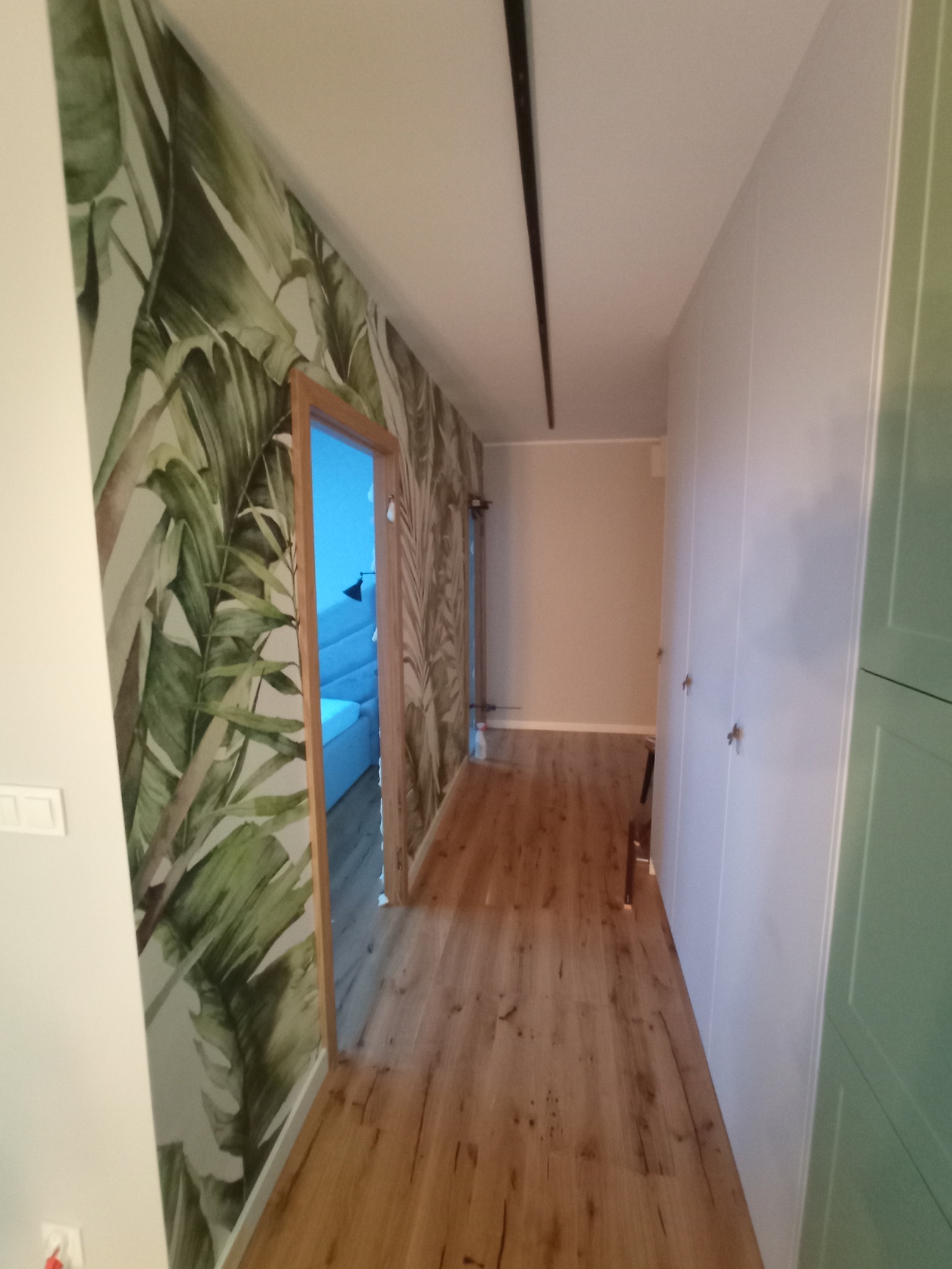 Korytarz z drewnianą podłogą i szafą w zabudowie, ściana z fototapetą w motywy roślinne, widok na sypialnię z niebieskim łóżkiem przez otwarte drzwi z drewnianą ościeżnicą.