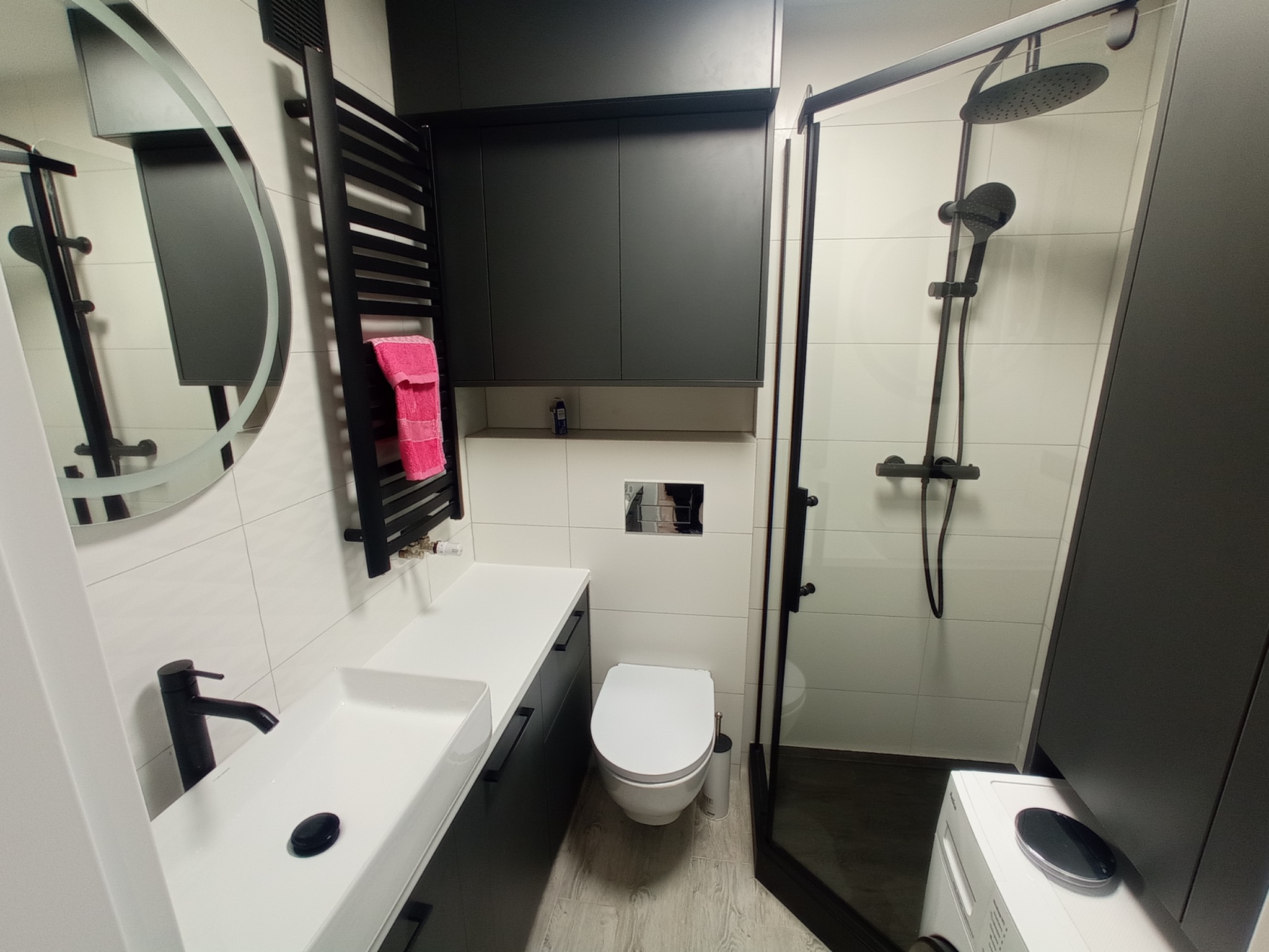 Nowoczesna łazienka z czarnymi akcentami: armatura, grzejnik drabinkowy z różowym ręcznikiem, kabina prysznicowa typu walk-in z deszczownicą i szafki wiszące nad toaletą.