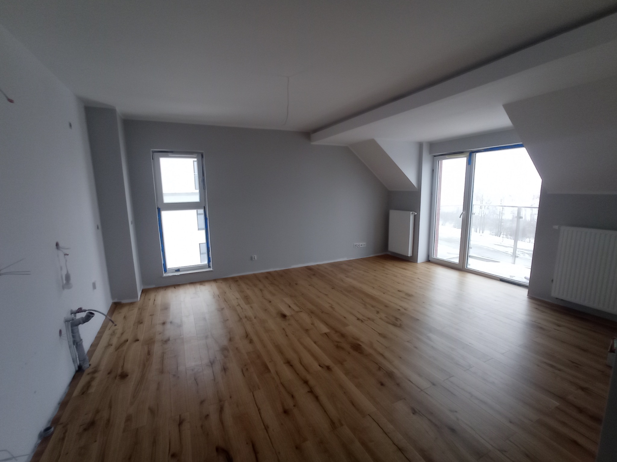 Przestronne, jasne poddasze po remoncie z panelami podłogowymi w odcieniach brązu, szare ściany, białe okna i grzejniki. Widoczne wystające rury instalacji na ścianie w lewym dolnym rogu.