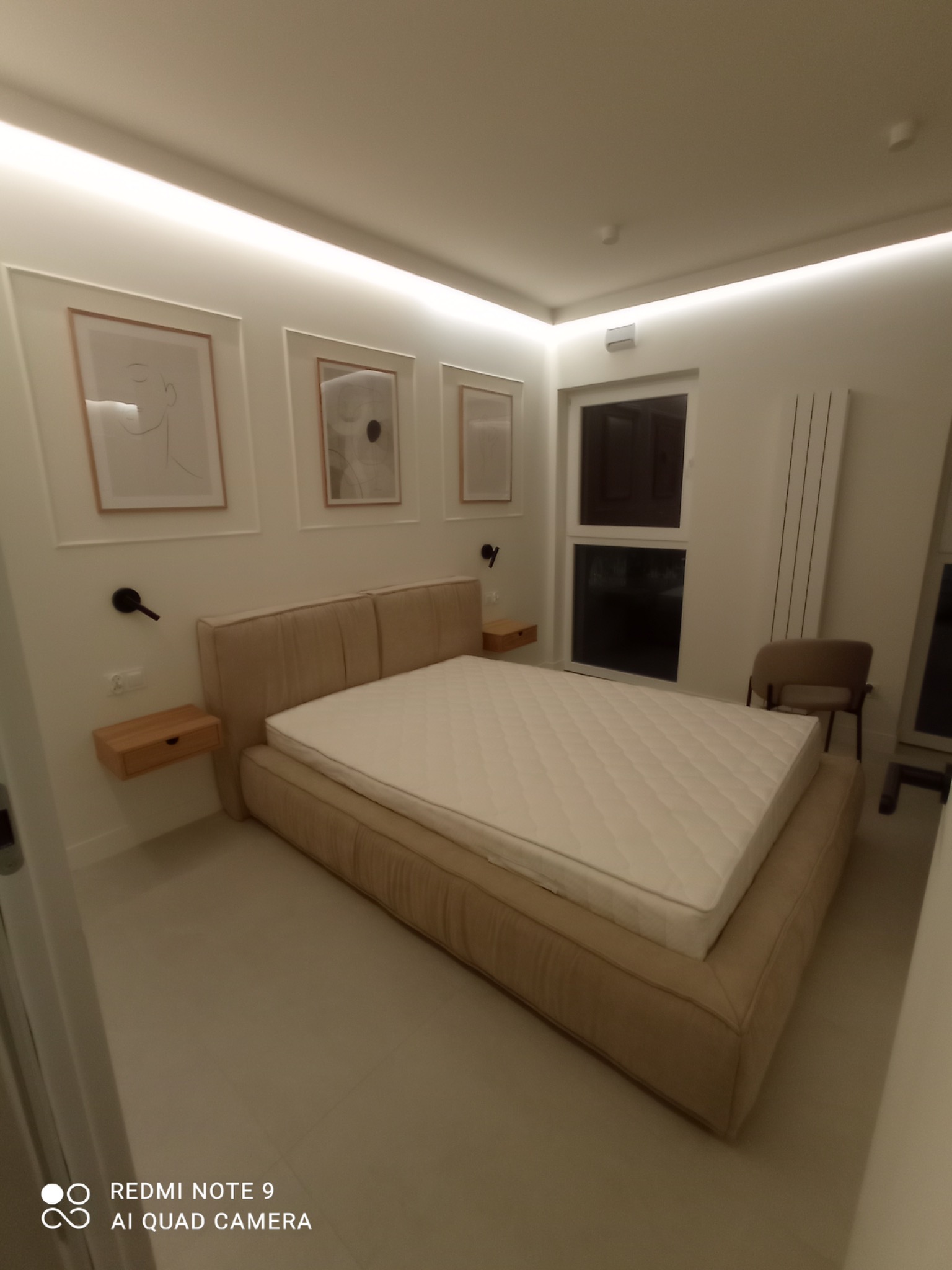 Nowoczesna sypialnia w neutralnych barwach z beżowym łóżkiem kontynentalnym, drewnianymi szafkami nocnymi i minimalistycznymi obrazami na ścianie, oświetlona listwami LED.