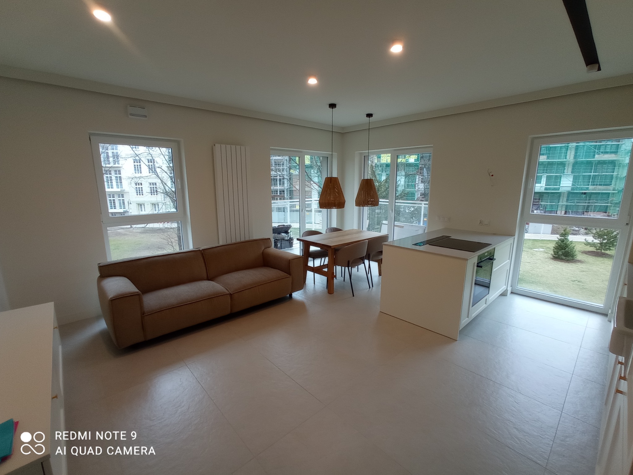 Jasne, przestronne wnętrze mieszkania po remoncie: beżowa sofa, drewniany stół z krzesłami, biała wyspa kuchenna, duże okna z widokiem na zewnątrz. Minimalistyczny design z lampami wiszącymi...