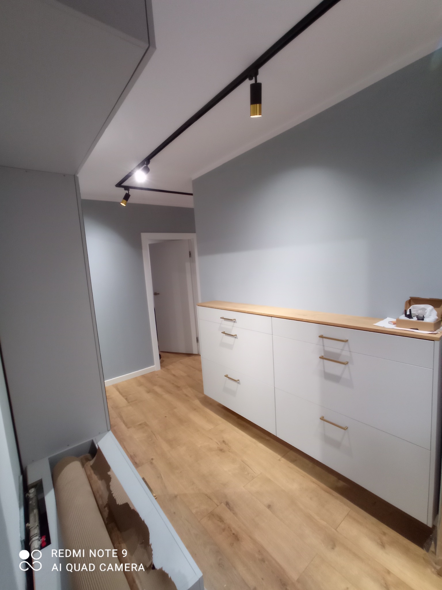 Korytarz po odświeżeniu: szare ściany, jasna podłoga z paneli, biała komoda z drewnianym blatem i złotymi uchwytami, oświetlenie szynowe z czarnymi reflektorami.