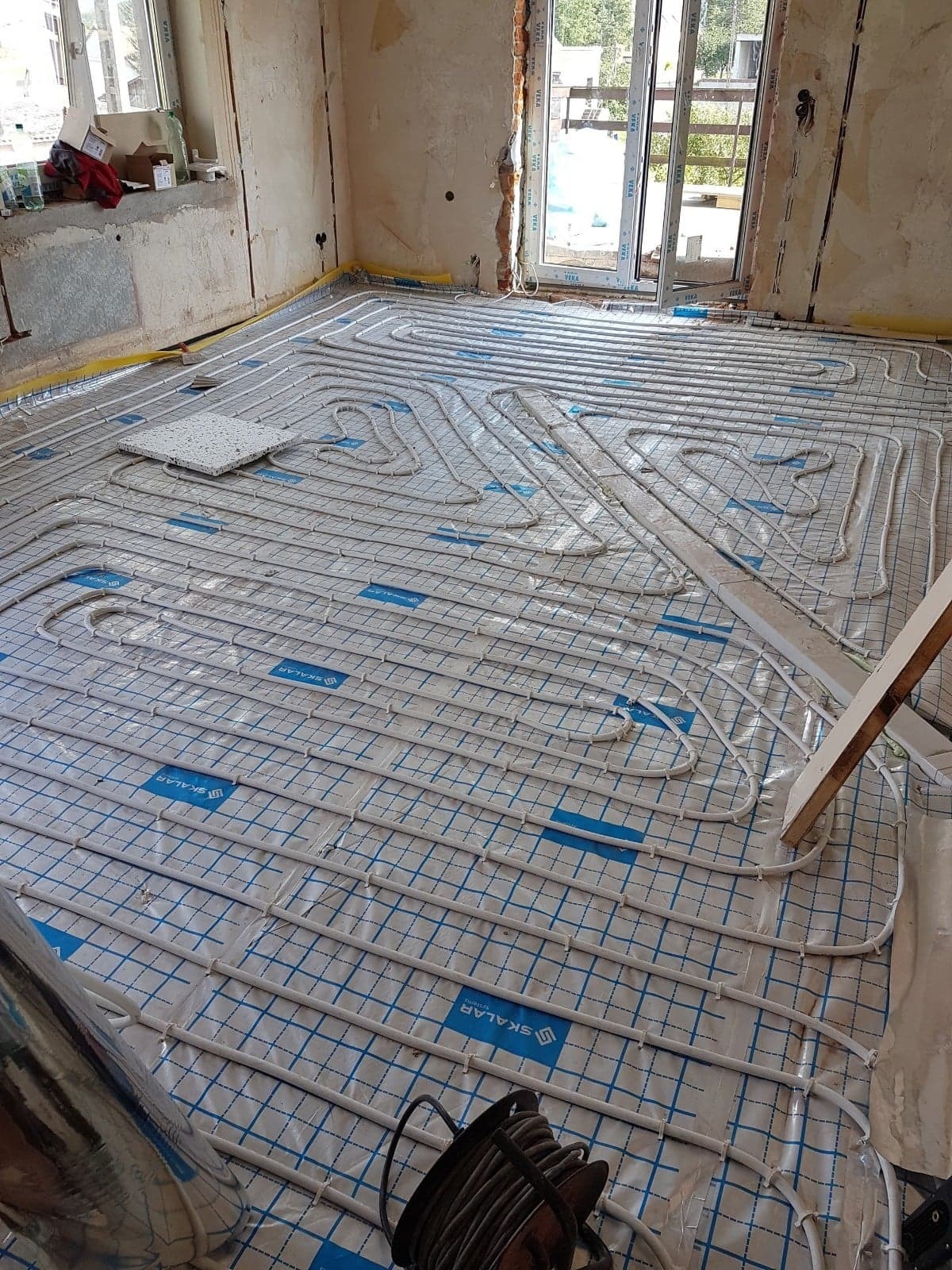 Rozłożona instalacja ogrzewania podłogowego z rurkami na folii z nadrukiem siatki i logo 'SKALAR', widoczne elementy montażowe i narzędzia w surowym pomieszczeniu.