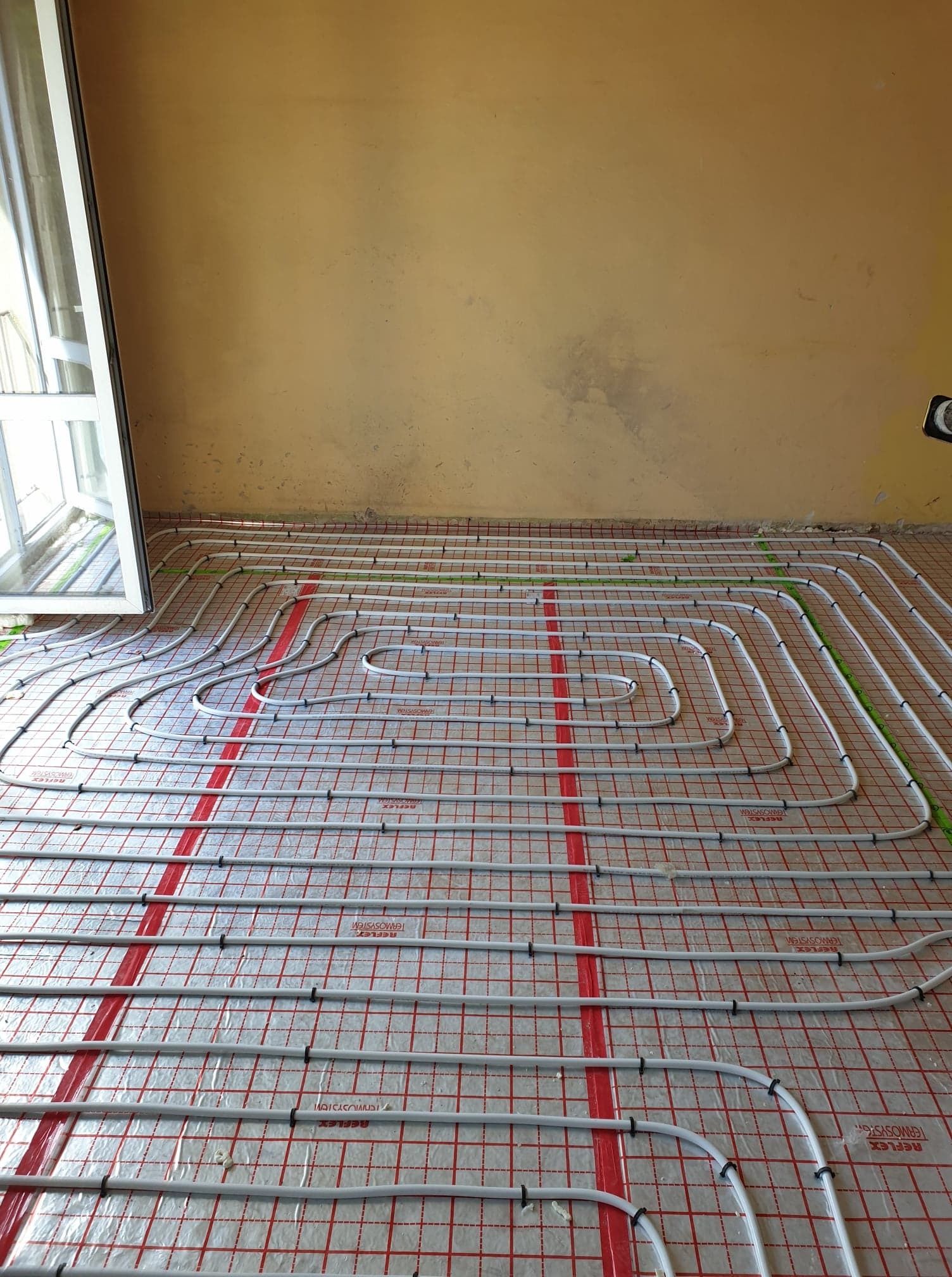 Rozłożone rury ogrzewania podłogowego na folii z czerwoną siatką, widoczne czarne uchwyty mocujące rury, zielone rurki na obrzeżach, ściana w tle w odcieniach beżu, okno po lewej stronie.