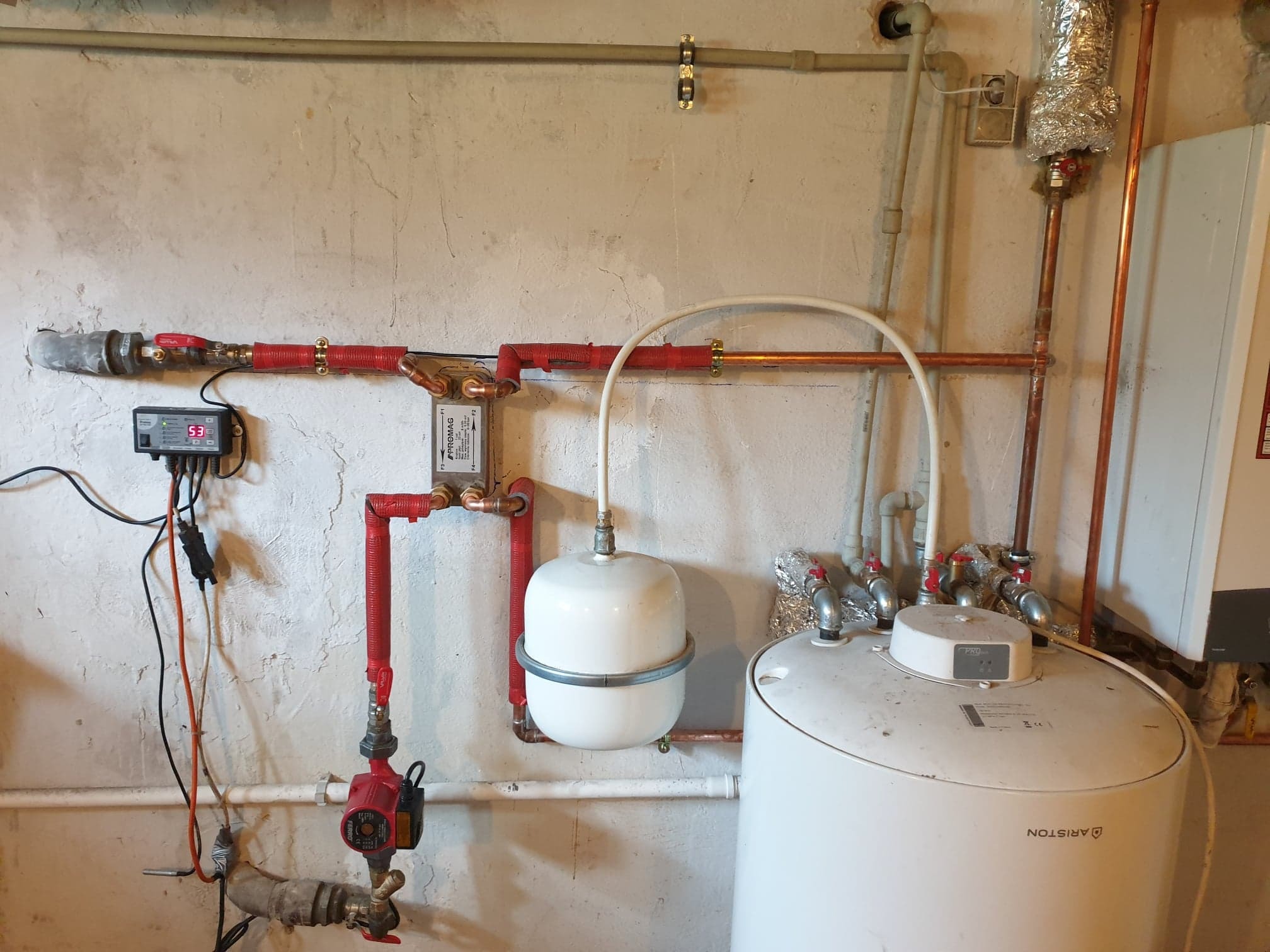 Instalacja grzewcza w piwnicy: biały zbiornik Ariston, czerwone rury miedziane z izolacją, pompa obiegowa Ferro, sterownik elektroniczny na ścianie otynkowanej na biało.