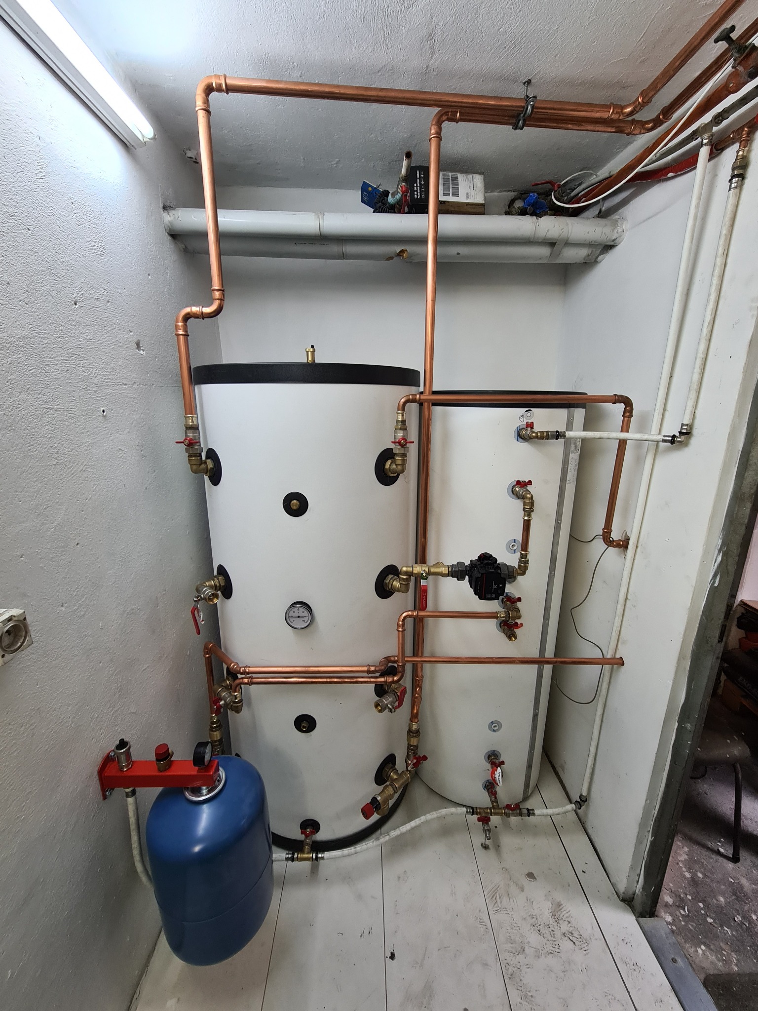 Dwa białe zbiorniki buforowe ciepła w kotłowni, połączone miedzianymi rurami, z widocznym manometrem i niebieskim naczyniem przeponowym na ścianie.
