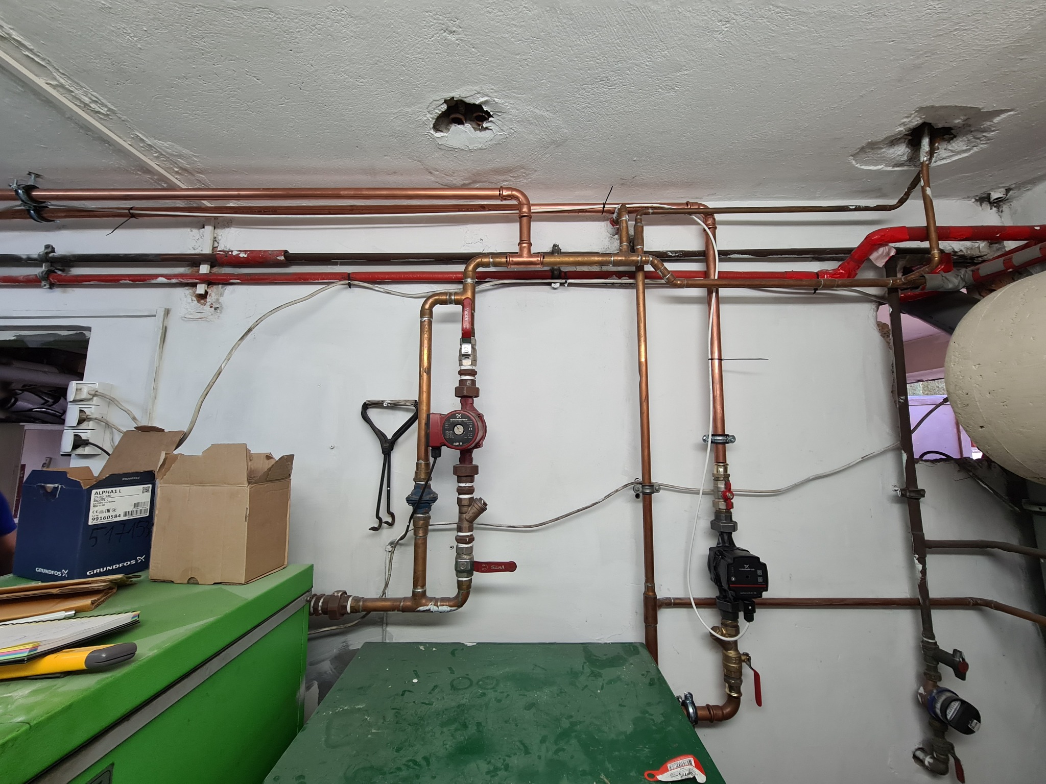 Widok na fragment instalacji grzewczej z miedzianymi rurami, pompami obiegowymi Grundfos, zaworami oraz uszkodzeniami w suficie. Na pierwszym planie widoczne kartony po pompach i zielony blat.