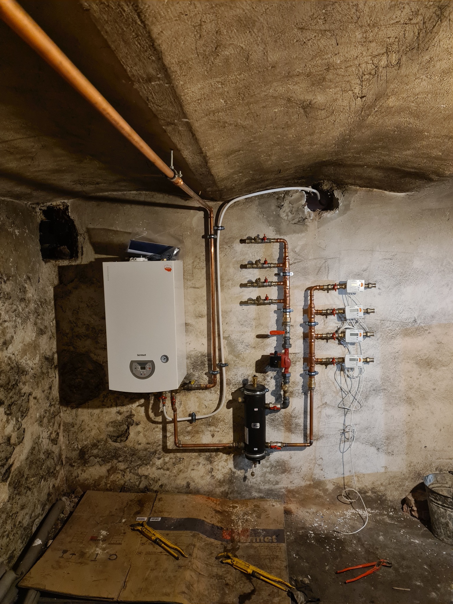 Nowo zamontowany piec gazowy Termet z rozbudowaną instalacją hydrauliczną, w tym rozdzielacz z zaworami i liczniki ciepła, na tle surowej ściany piwnicznej z widocznymi rurami miedzianymi...