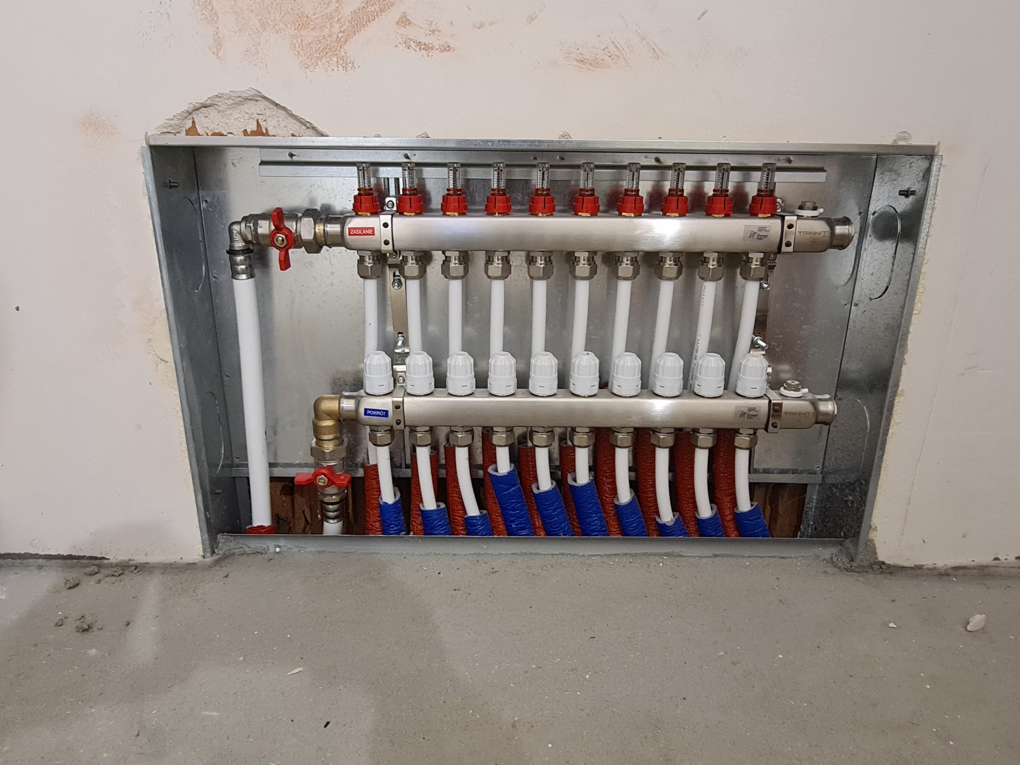 Rozdzielacz ogrzewania podłogowego zamontowany w metalowej szafce w ścianie, z widocznymi rurami doprowadzającymi wodę w izolacji czerwonej i niebieskiej.