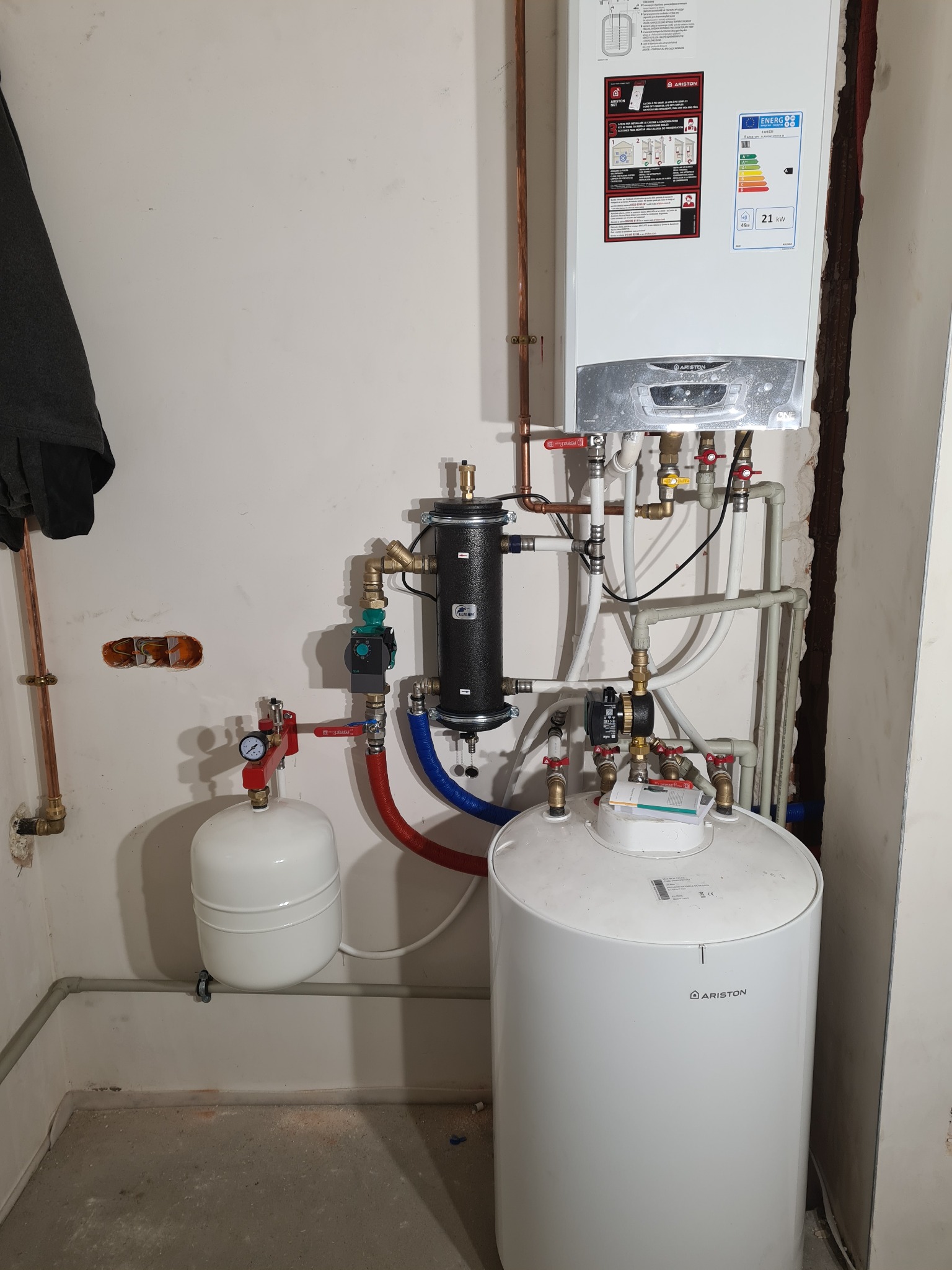 Kocioł gazowy Ariston z podłączonym zasobnikiem wody i elementami instalacji, widoczne rury miedziane i plastikowe, manometr z czerwoną wskazówką, ściana z widocznymi śladami po instalacji.