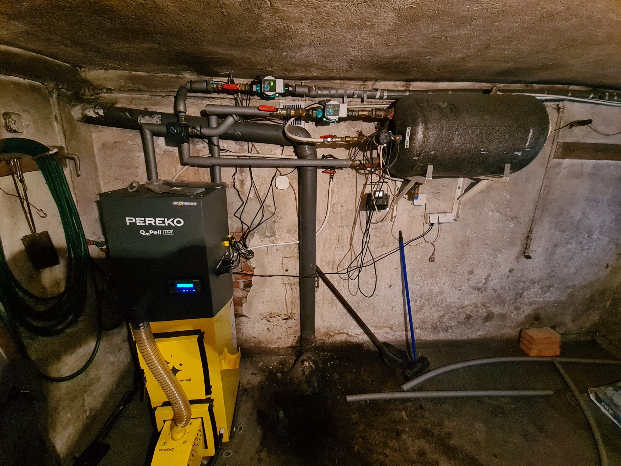 Instalacja kotła na pellet Pereko w piwnicy, widoczne rury, zbiornik buforowy i elementy instalacji grzewczej w surowym pomieszczeniu.