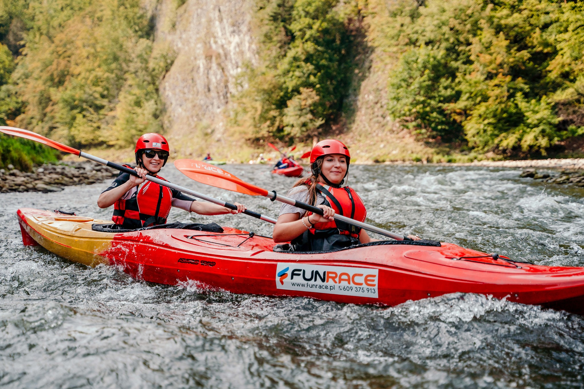 Dwie osoby w czerwonych kaskach i kamizelkach asekuracyjnych płyną kajakiem po rwącej rzece w otoczeniu zielonych drzew i skalistych brzegów. Kajak jest czerwono-żółty z naklejką 'FUNRACE'.