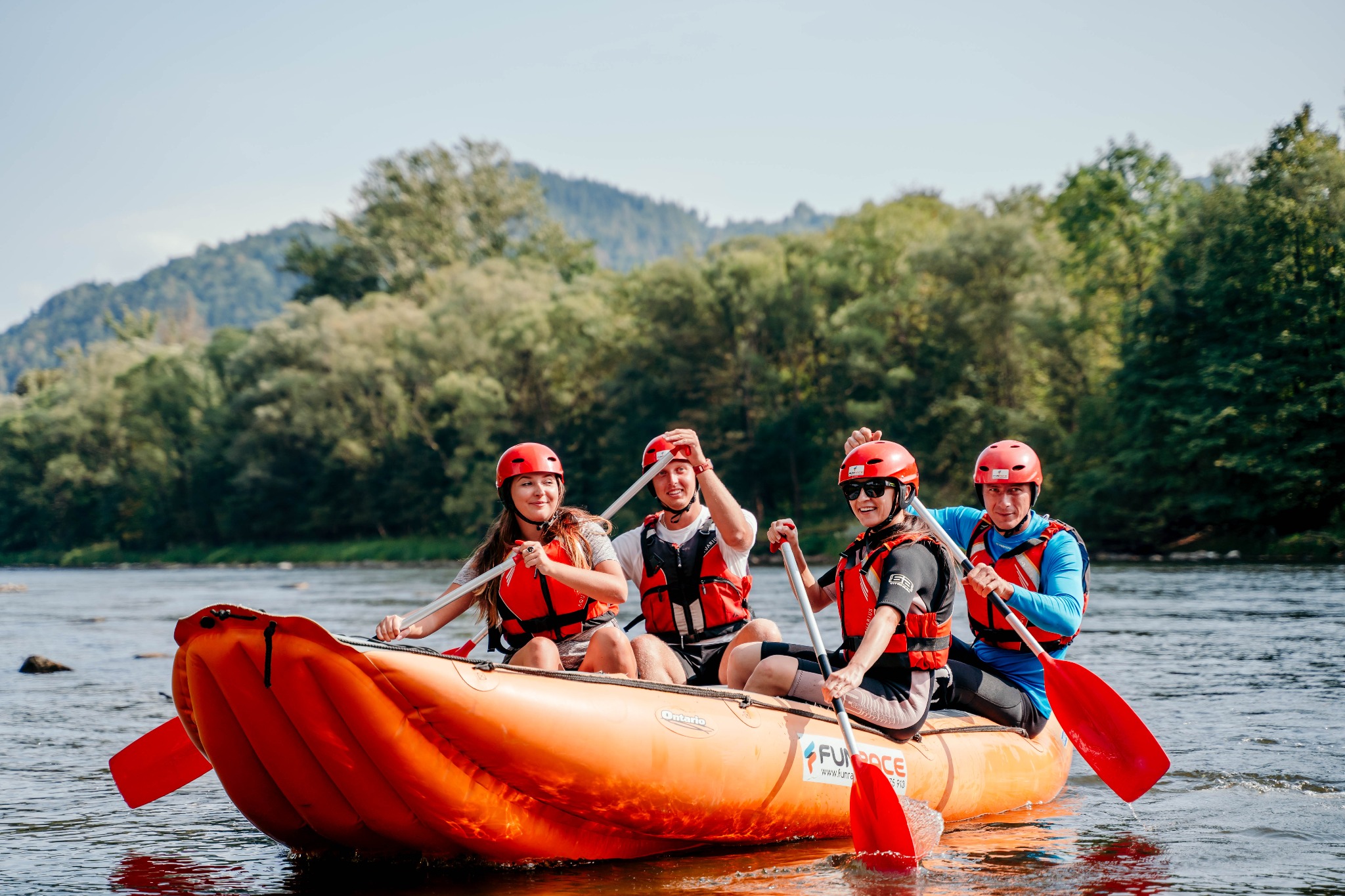 Grupa czterech osób w czerwonych kaskach i kamizelkach ratunkowych płynie pomarańczowym pontonem po rzece, trzymając wiosła, w tle zalesione wzgórza.